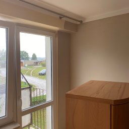 Grzegorz Sikora - Pokój z otwartym oknem balkonowym w trakcie odświeżania, ściany pomalowane na beżowo, widoczny fragment drewnianej szafki i niedomalowany fragment pod sufitem.