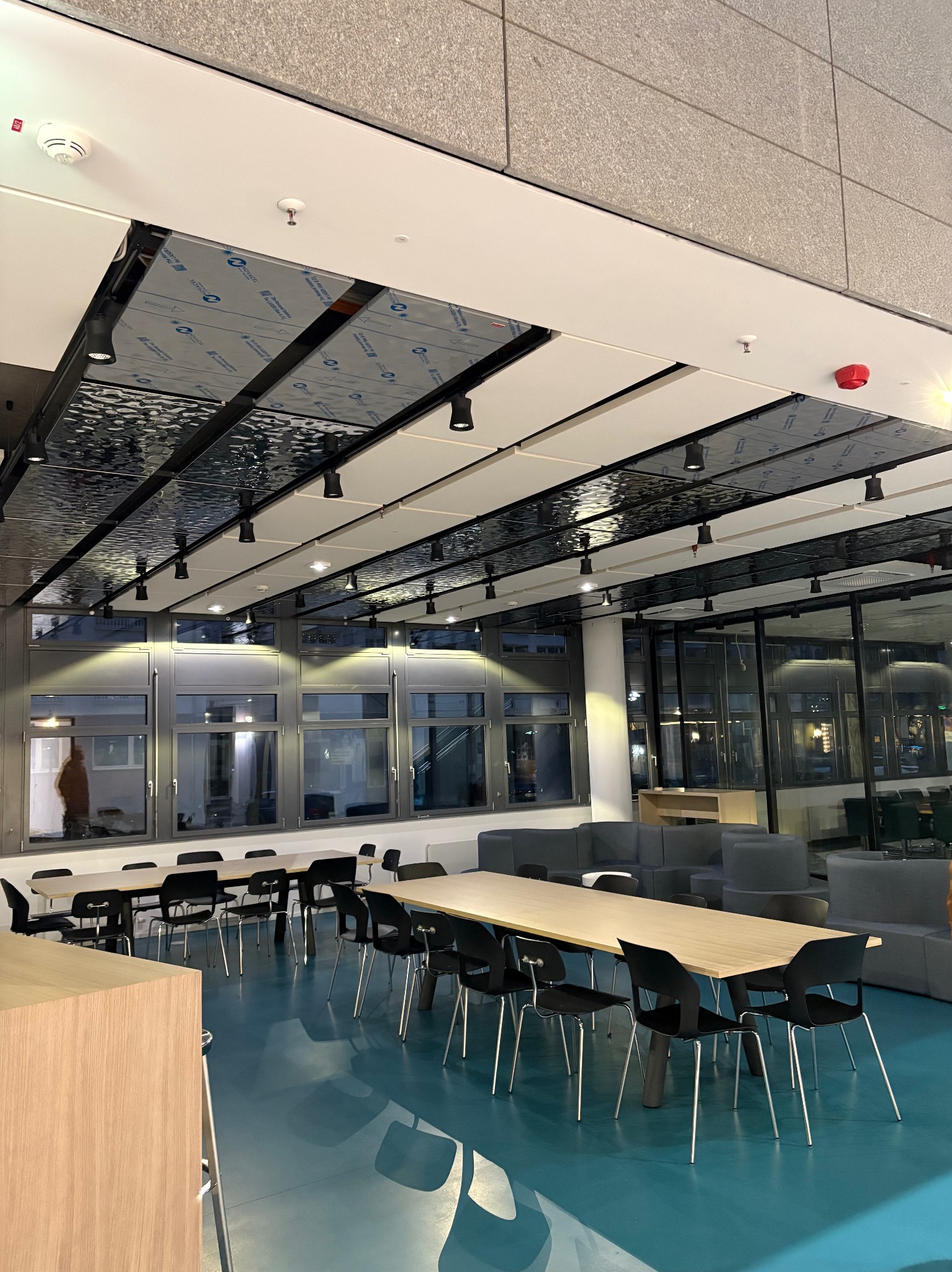 Nowoczesne wnętrze biurowe z długimi stołami, czarnymi krzesłami, niebieską podłogą i sufitem z elementami metalowymi oraz oświetleniem punktowym.