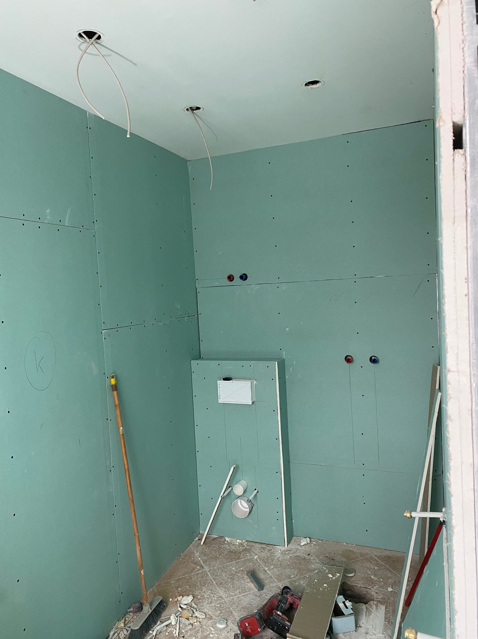 Łazienka w trakcie montażu płyt kartonowo-gipsowych, widoczne otwory na oświetlenie w suficie, instalacja wodna przygotowana pod armaturę, na podłodze resztki materiałów i narzędzia.