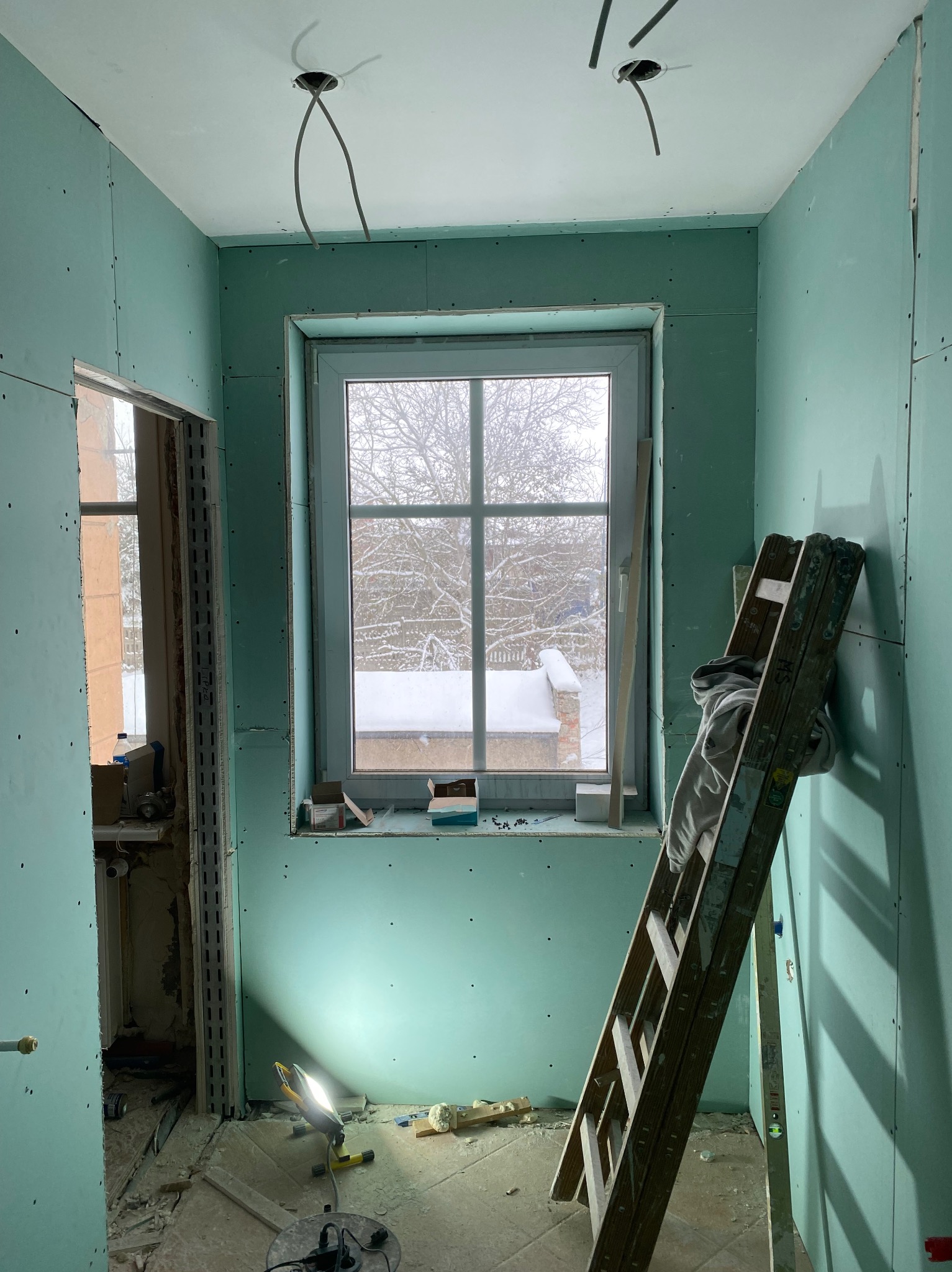 Wnętrze w trakcie montażu płyt gipsowo-kartonowych, widoczne ściany z płyt, drabina oparta o ścianę, okno z widokiem na zaśnieżone drzewa oraz lampa warsztatowa oświetlająca podłogę.