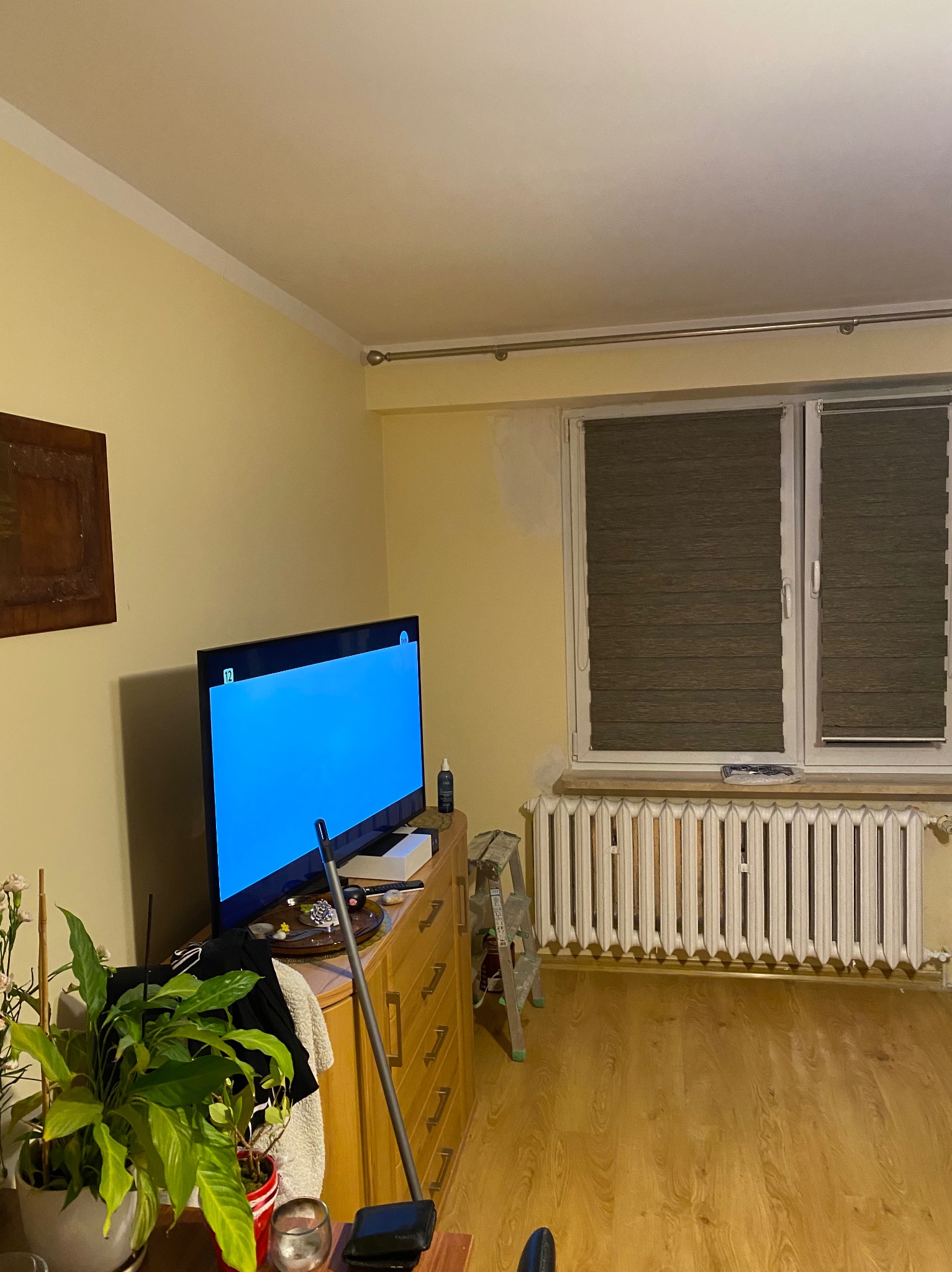 Pokój z żółtymi ścianami, grzejnikiem i oknem z żaluzjami, fragment ściany przygotowany do malowania, drabina oparta o ścianę, meble zagracone przedmiotami, w rogu kwiat doniczkowy.