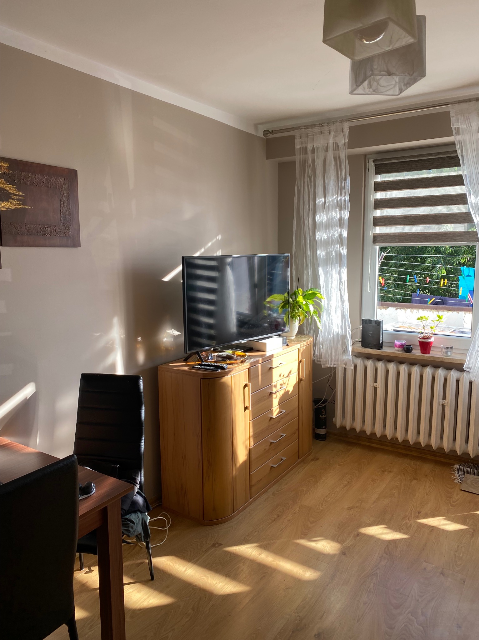 Pokój dzienny z drewnianą komodą pod telewizorem, drewnianą podłogą i grzejnikiem pod oknem, na którym suszą się ubrania.