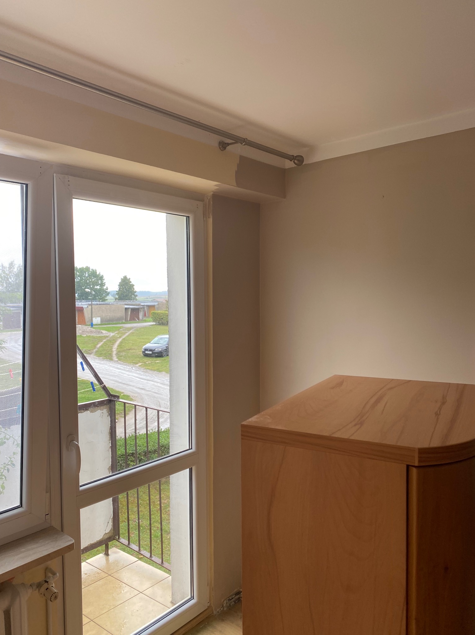 Pokój z otwartym oknem balkonowym w trakcie odświeżania, ściany pomalowane na beżowo, widoczny fragment drewnianej szafki i niedomalowany fragment pod sufitem.
