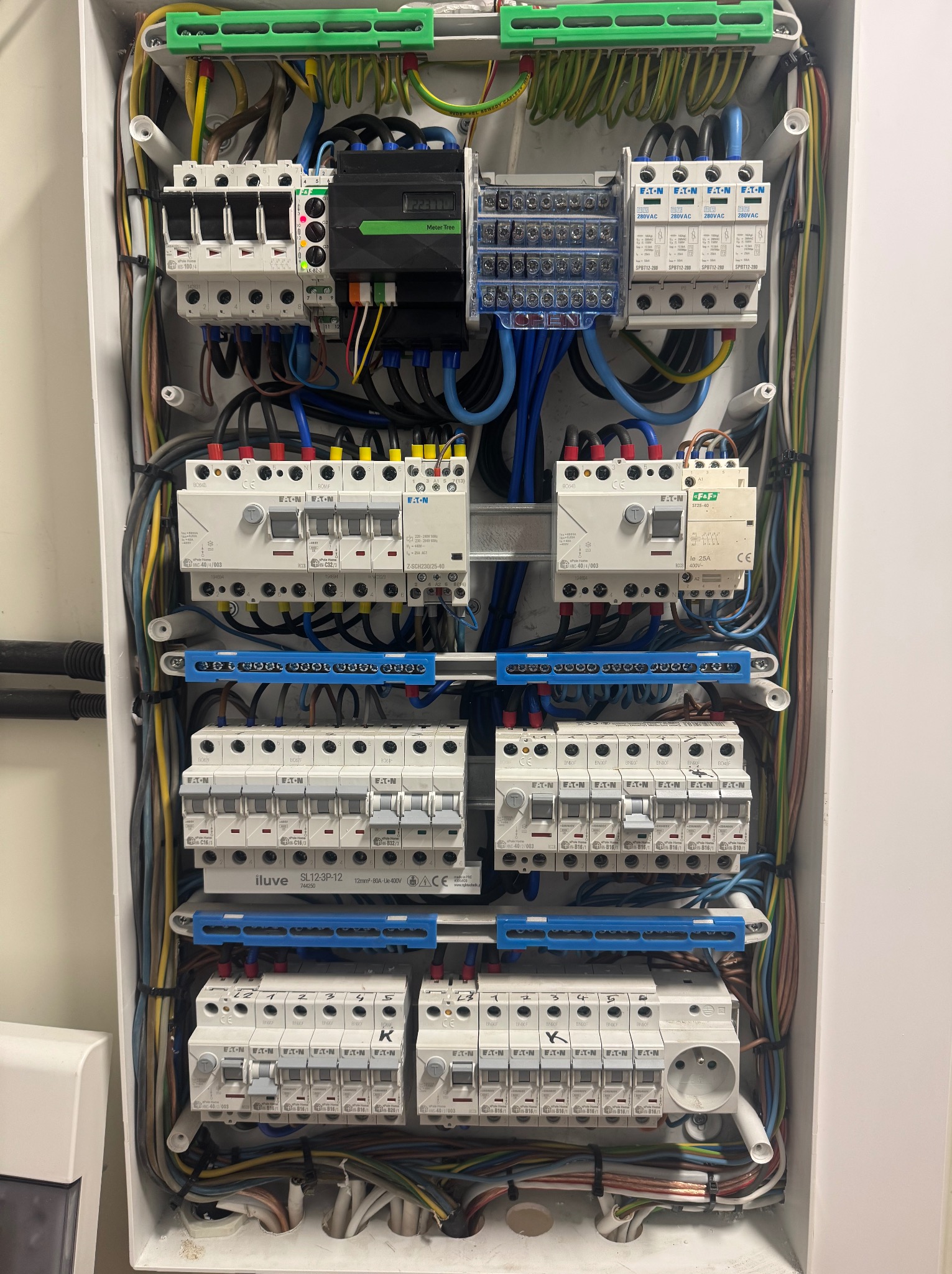 Rozdzielnica elektryczna z zainstalowanymi bezpiecznikami, przewodami i modułem pomiarowym. Widoczne elementy marki Eaton i Iluve. Instalacja w białej obudowie.