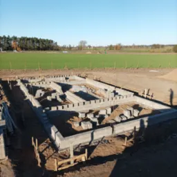 Budowa fundamentów domu jednorodzinnego z bloczków betonowych, widok z góry na częściowo ukończoną konstrukcję na tle zielonego pola i drzew