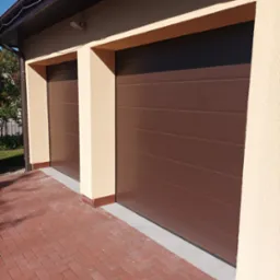 Dwa brązowe garaże segmentowe z elewacją w kolorze kremowym i kostką brukową w jodełkę.