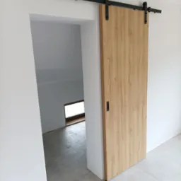 Drewniane drzwi przesuwne w stylu rustykalnym na czarnej szynie, zamontowane w otworze drzwiowym, z widokiem na surowe wnętrze z oknem.