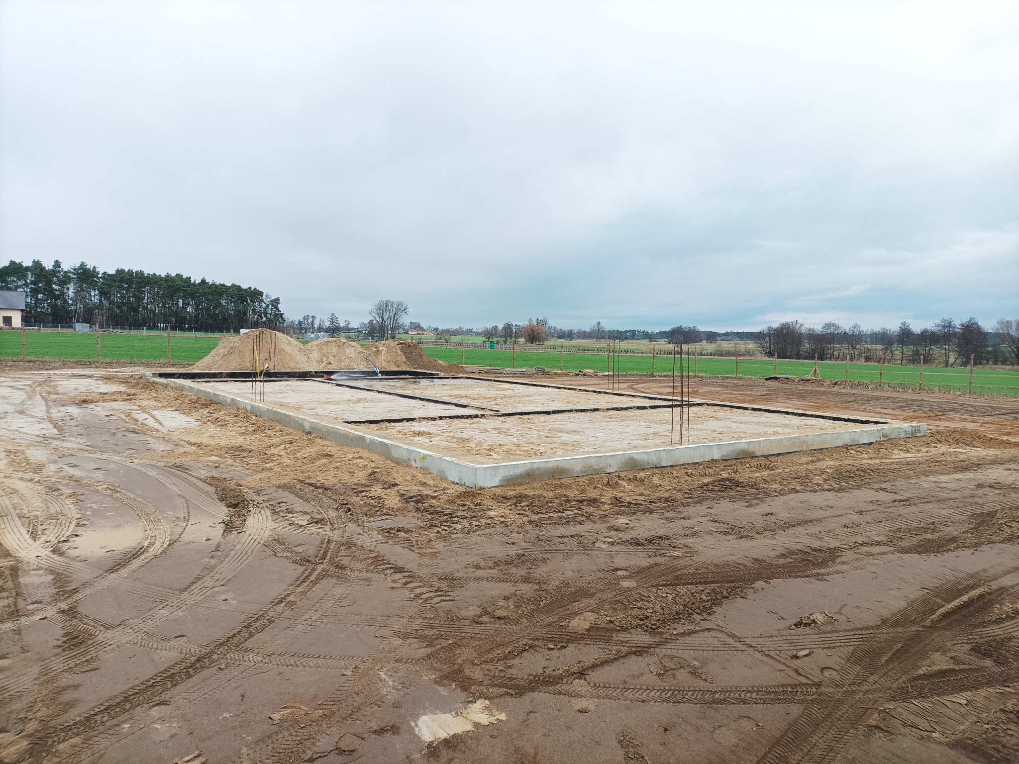 Zalany betonowy fundament z wystającymi prętami zbrojeniowymi na piaszczystym terenie, widoczne ślady opon maszyn budowlanych, w tle łąka i drzewa pod pochmurnym niebem.