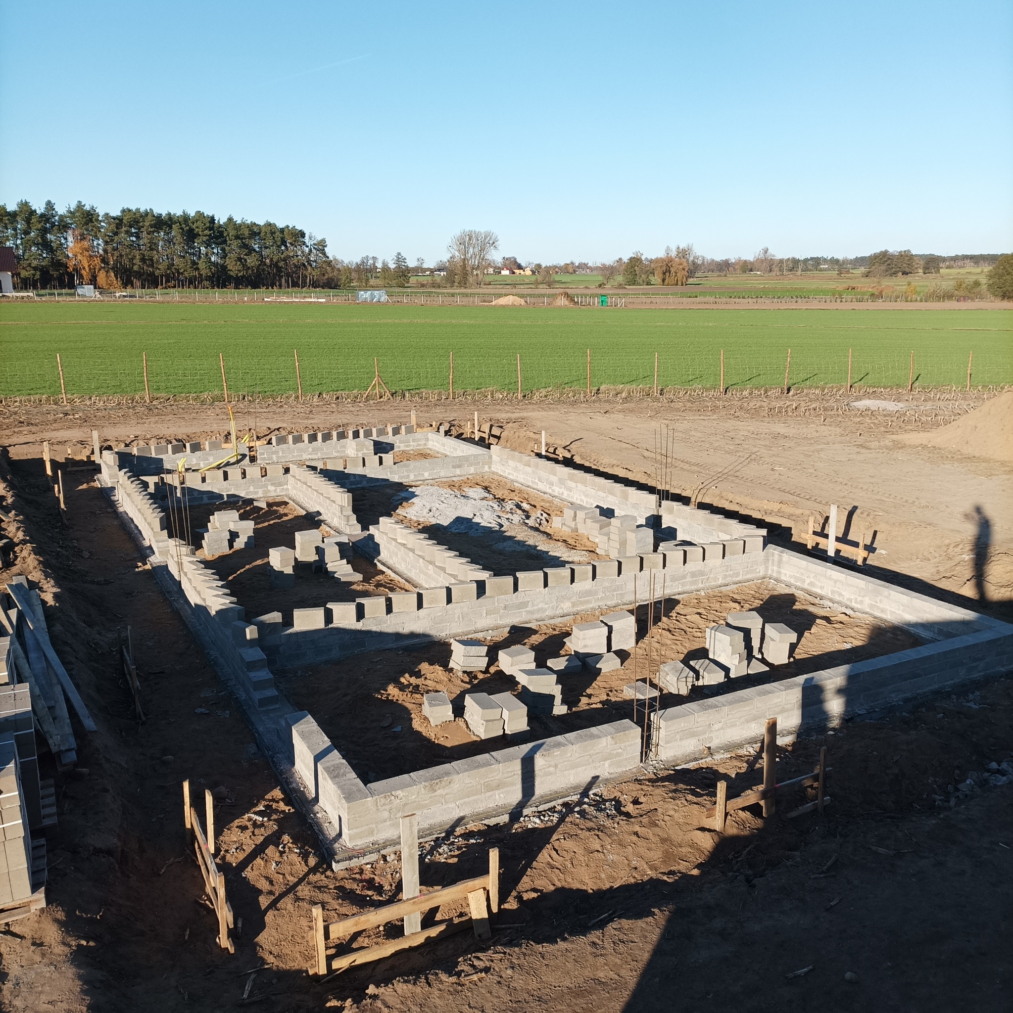 Budowa fundamentów domu jednorodzinnego z bloczków betonowych, widok z góry na częściowo ukończoną konstrukcję na tle zielonego pola i drzew