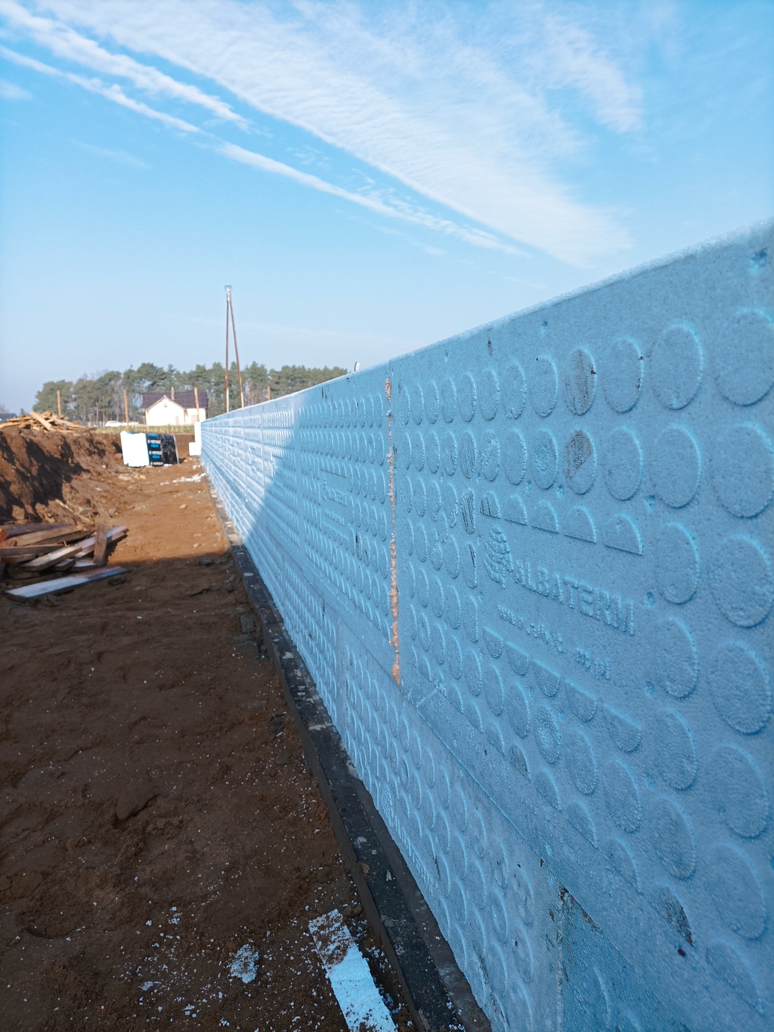 Nisko położony, szeroki kadr na niebieską izolację fundamentów z widocznym logo 'ALBATERM' i okrągłymi wytłoczeniami, na tle placu budowy i błękitnego nieba.