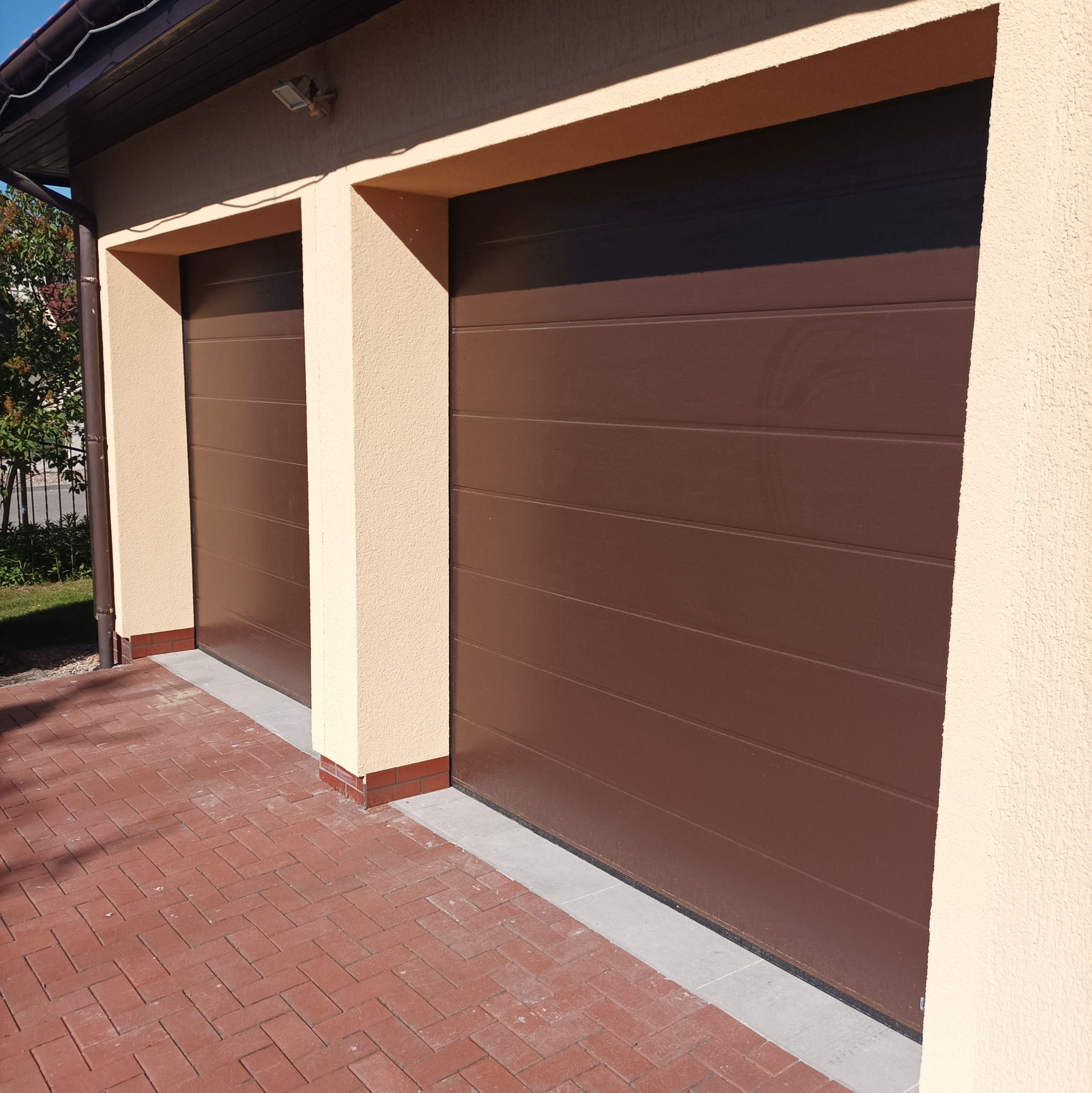 Dwa brązowe garaże segmentowe z elewacją w kolorze kremowym i kostką brukową w jodełkę.