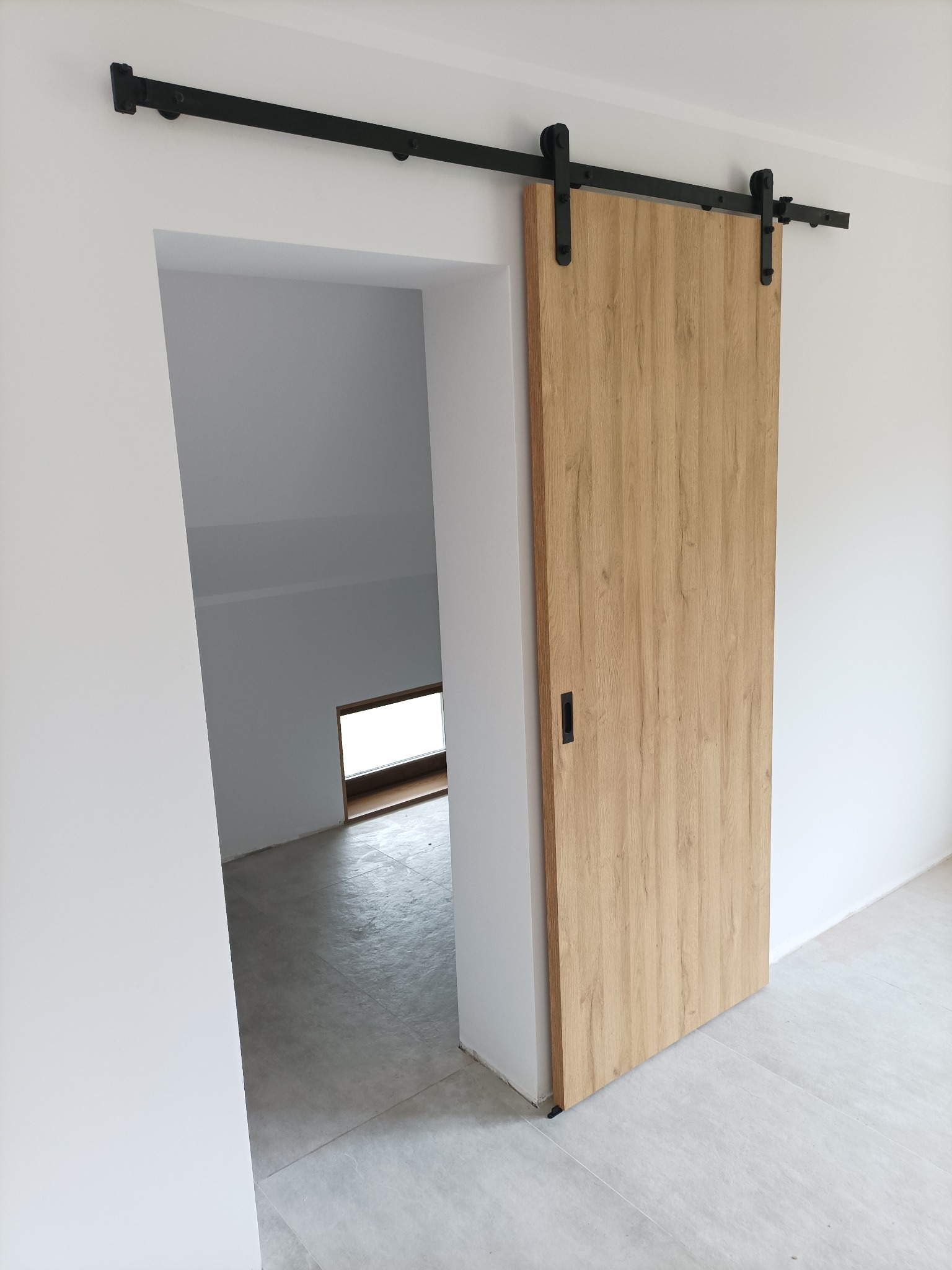 Drewniane drzwi przesuwne w stylu rustykalnym na czarnej szynie, zamontowane w otworze drzwiowym, z widokiem na surowe wnętrze z oknem.