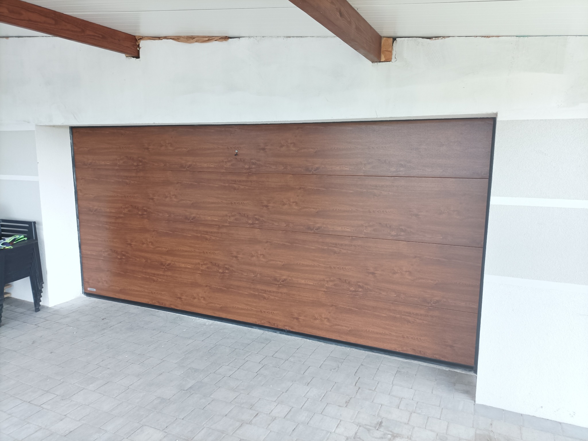 Zewnętrzna brama garażowa segmentowa z paneli imitujących drewno, widoczna od strony podjazdu wyłożonego szarą kostką brukową.
