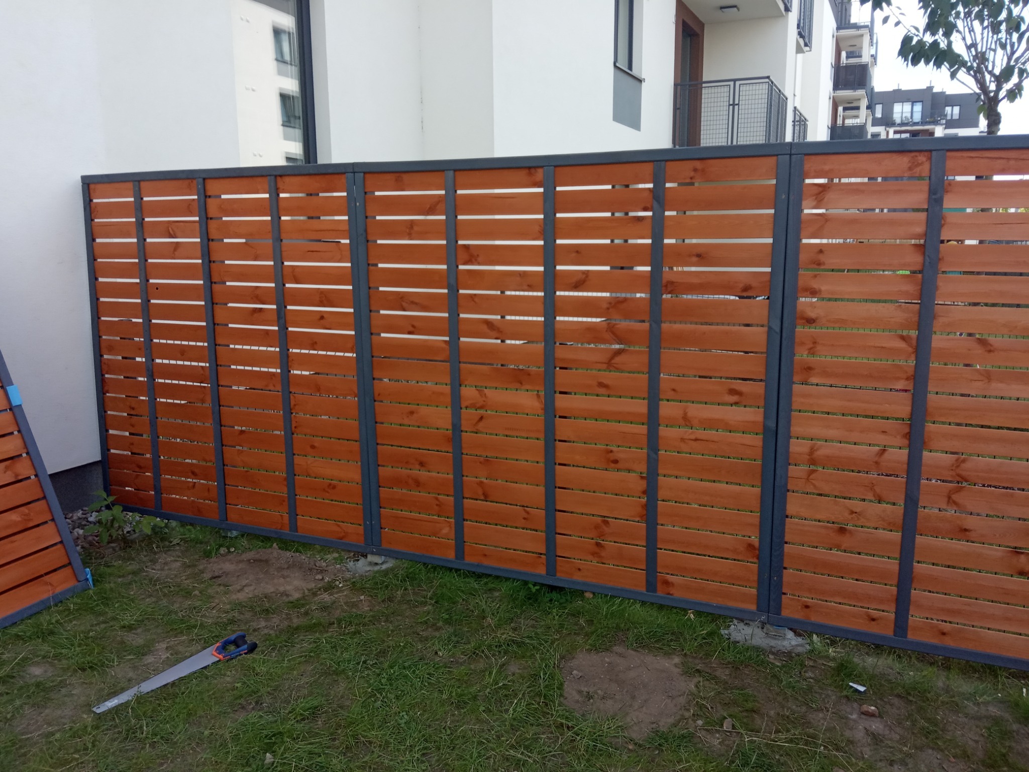 Nowoczesne, poziome ogrodzenie z drewnianych desek w kolorze ciepłego brązu, osadzone na szarej metalowej konstrukcji, widoczne na tle budynku mieszkalnego i trawnika, obok leży piła ręczna.