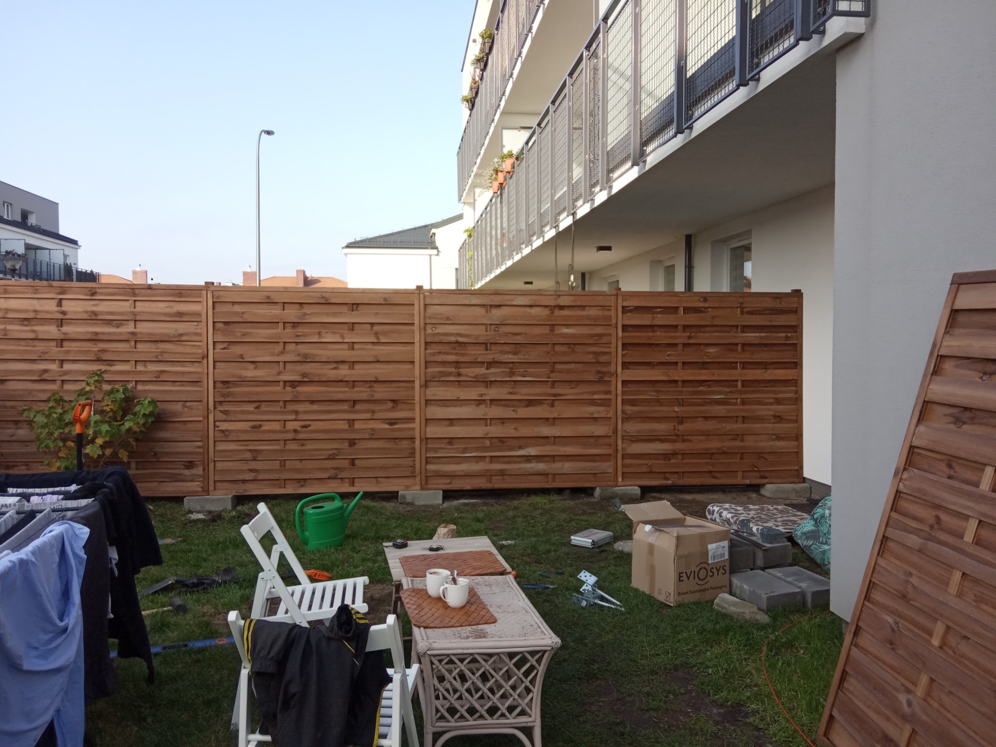 Nowo postawione, poziome, drewniane ogrodzenie panelowe oddzielające ogród od budynku wielorodzinnego, z widocznymi narzędziami, elementami montażowymi i dwoma kubkami z napojem na stoliku kawowym...