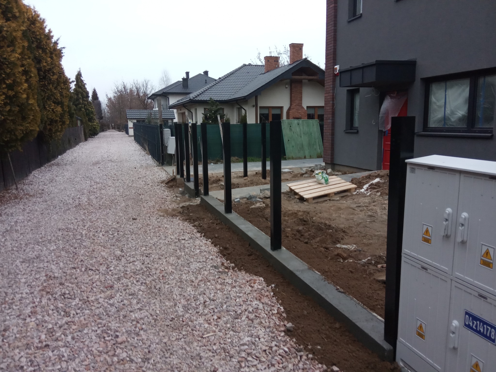 Wykop pod fundament ogrodzenia z widocznymi czarnymi słupkami w trakcie montażu, obok wysypany żwir, w tle zabudowa jednorodzinna.