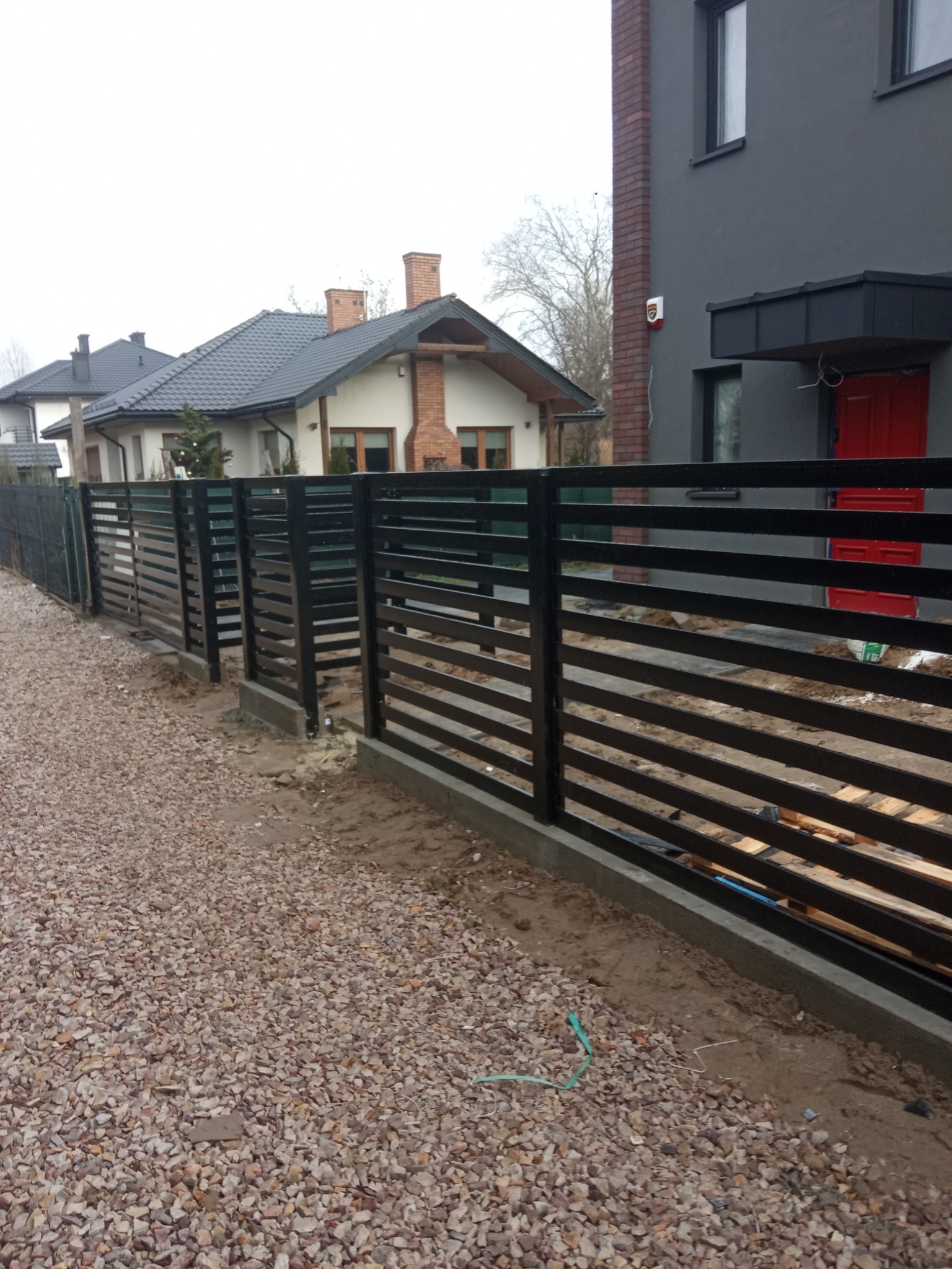 Nowoczesne, poziome ogrodzenie panelowe w kolorze czarnym, z widocznymi elementami montażowymi, na tle domów jednorodzinnych i żwirowego podłoża.
