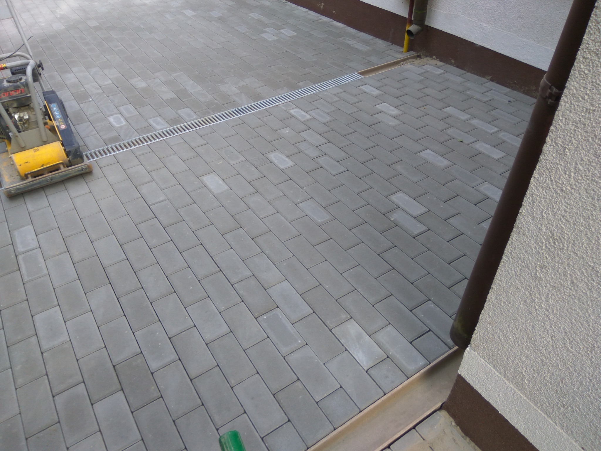 Ułożona szara kostka brukowa z odpływem liniowym przy ścianie budynku, ubijarka wibracyjna w rogu.
