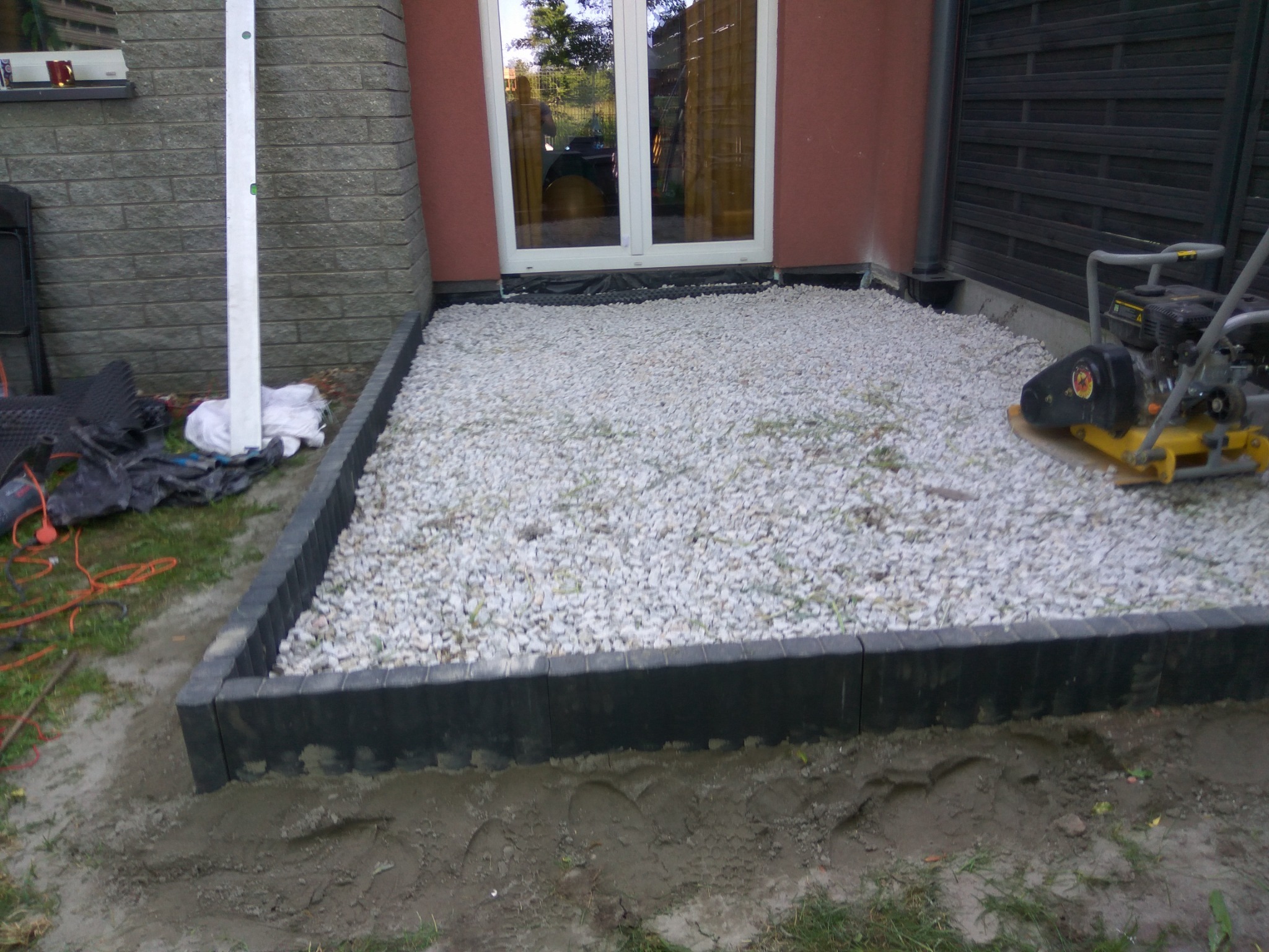 Przygotowany teren pod taras z obrzeżami z kostki, zasypany białym grysem, ubijarka w tle, widok na drzwi tarasowe.