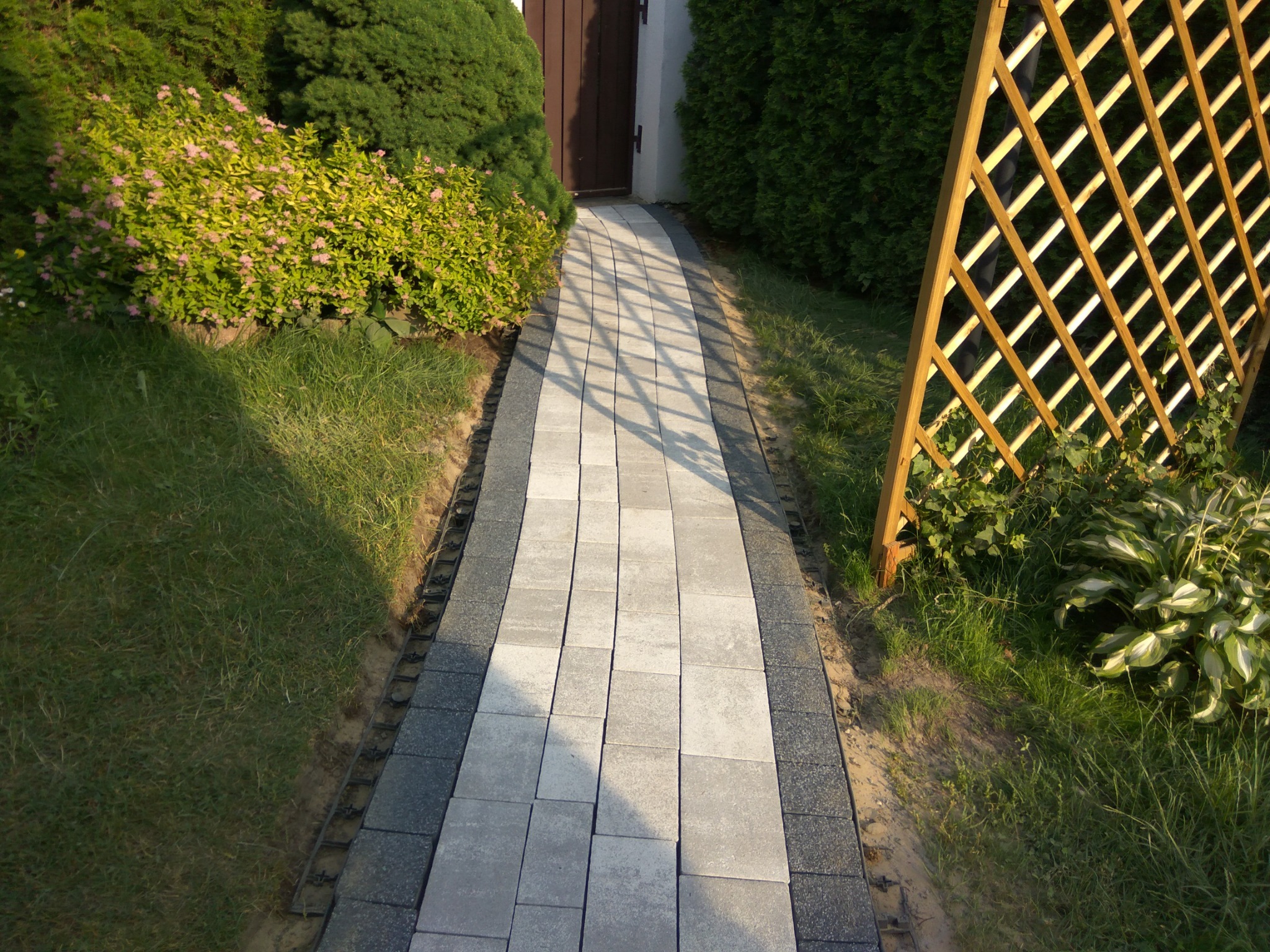 Ścieżka z jasnoszarej kostki brukowej z ciemnoszarym obramowaniem, prowadząca do drewnianych drzwi, otoczona zielenią i drewnianą kratownicą