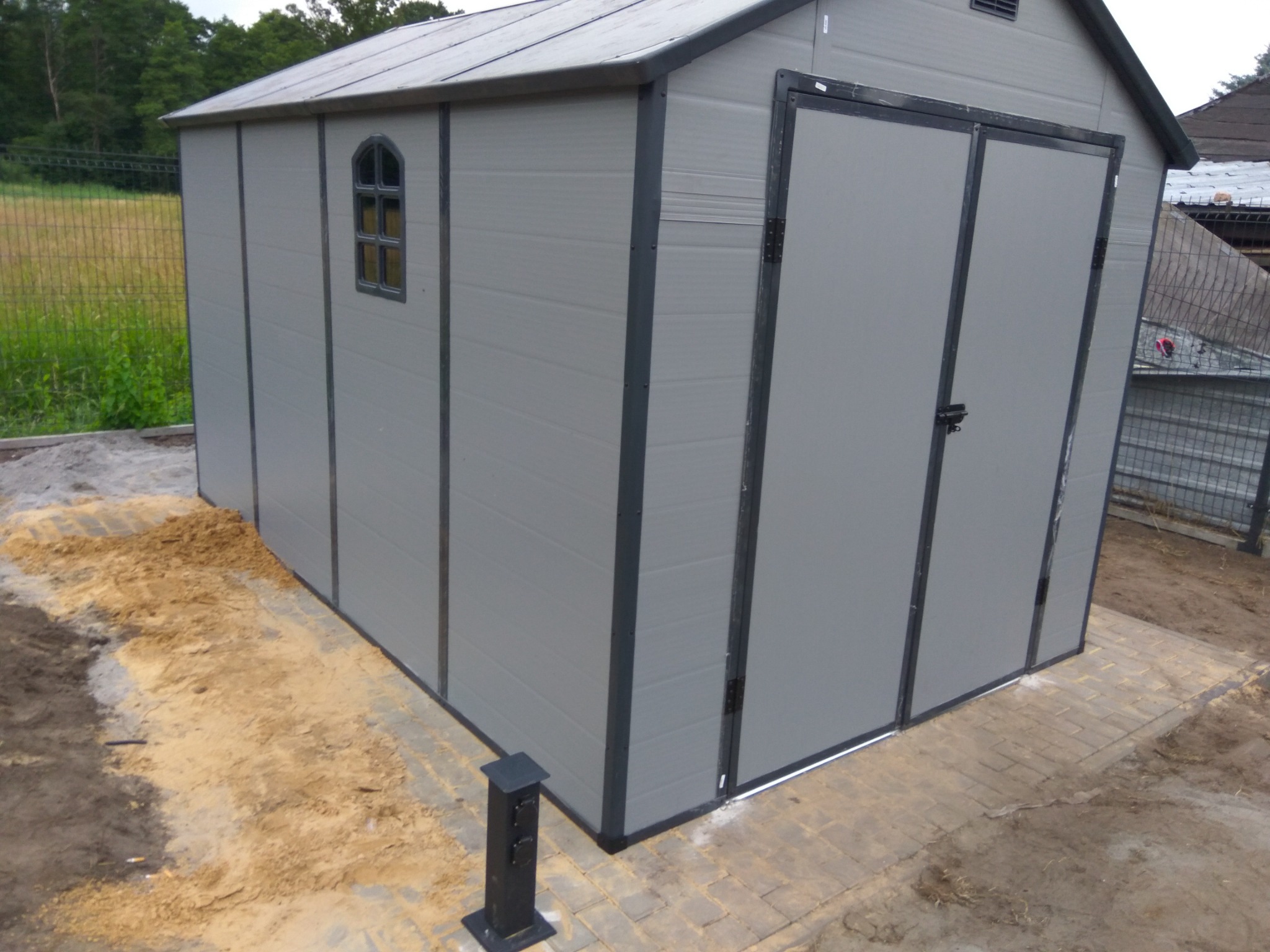 Szara, plastikowa szopa ogrodowa z małym okienkiem w kształcie łuku, stojąca na nowo ułożonej, brudnej kostce brukowej, obok gniazdka elektrycznego na zewnątrz.