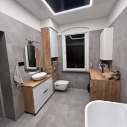 Wykończona łazienka w szarościach z drewnianymi akcentami, owalna umywalka na białej szafce z szufladami, okrągłe lustro, płytki imitujące beton na ścianach i podłodze, sufit z podświetleniem LED.