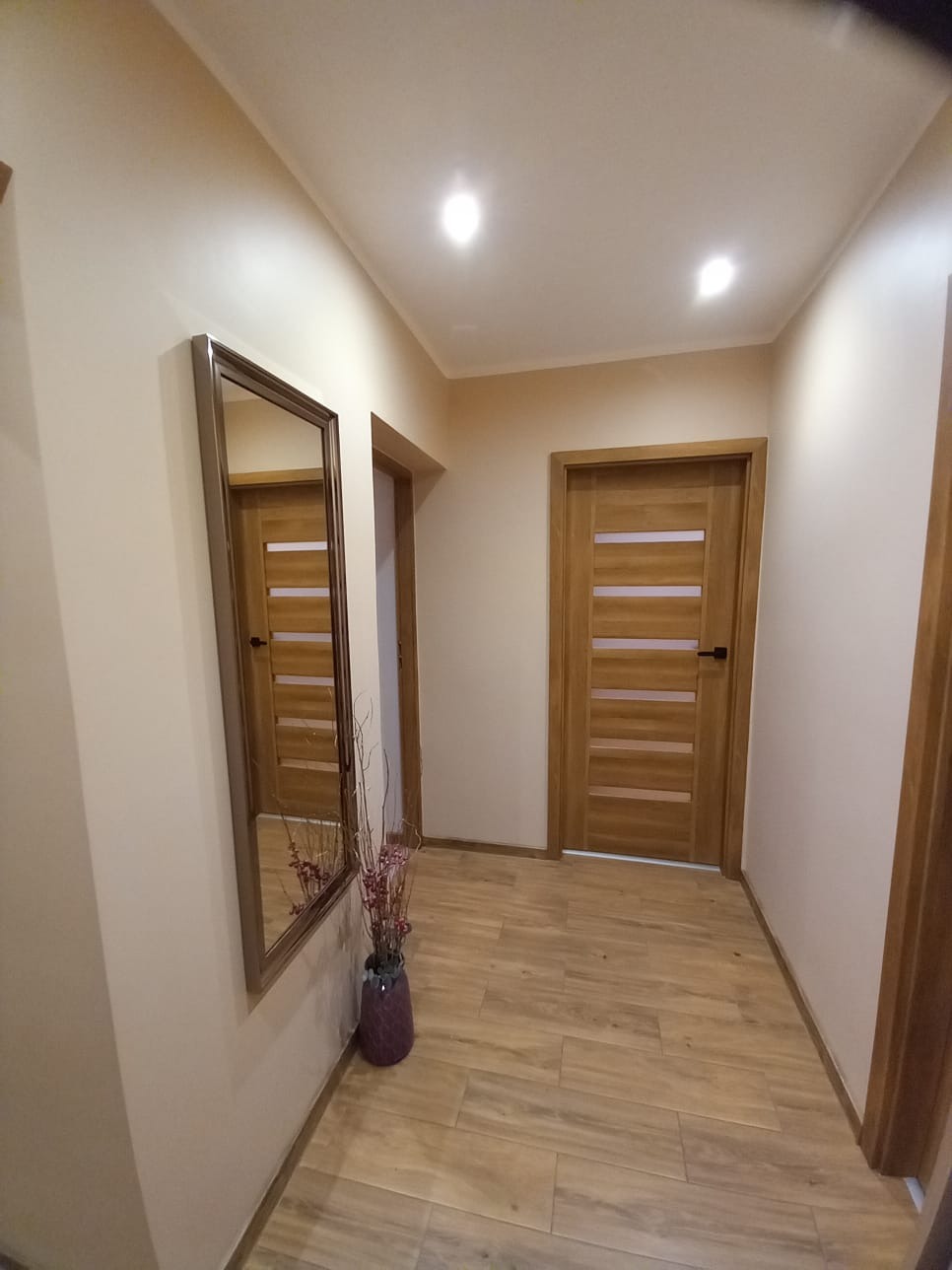 Korytarz po remoncie z drewnopodobną podłogą, lustrem w ramie i drewnianymi drzwiami z przeszkleniami, oświetlony trzema punktowymi lampami sufitowymi, z dekoracyjnym wazonem na podłodze.