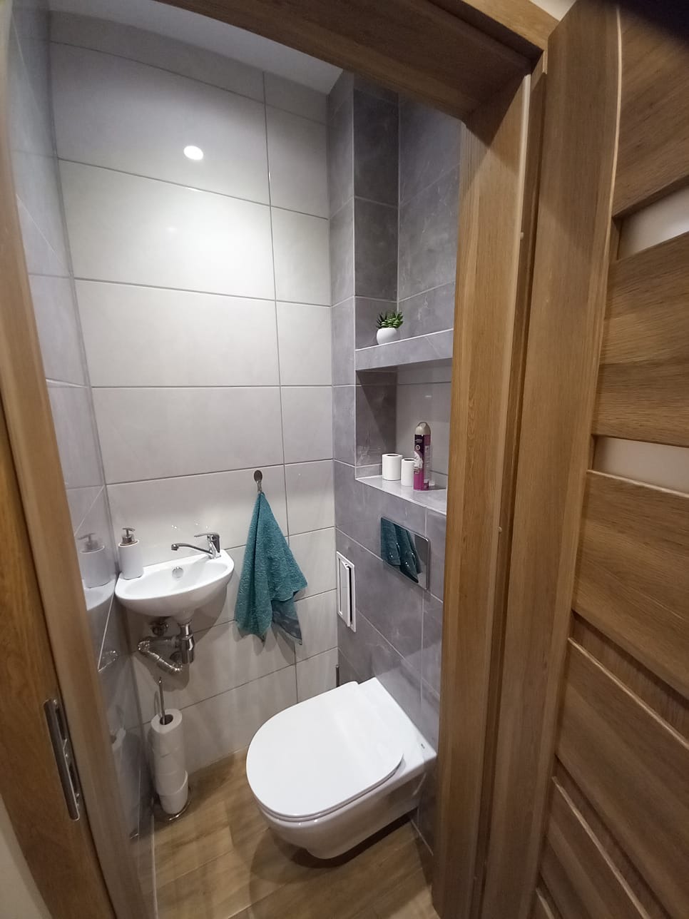 Mała, nowoczesna łazienka z wiszącą toaletą, umywalką narożną i półkami wnękowymi, wykończona szarymi płytkami w formacie kwadratu i drewnianymi drzwiami.