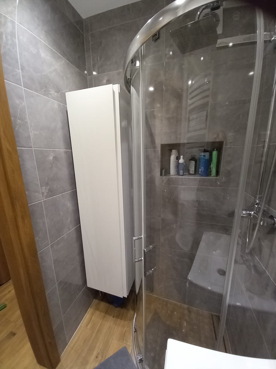 Nowoczesna łazienka z szarymi płytkami imitującymi marmur, kabiną prysznicową z deszczownicą, białą szafką wiszącą i drewnianą podłogą, widok z narożnika.