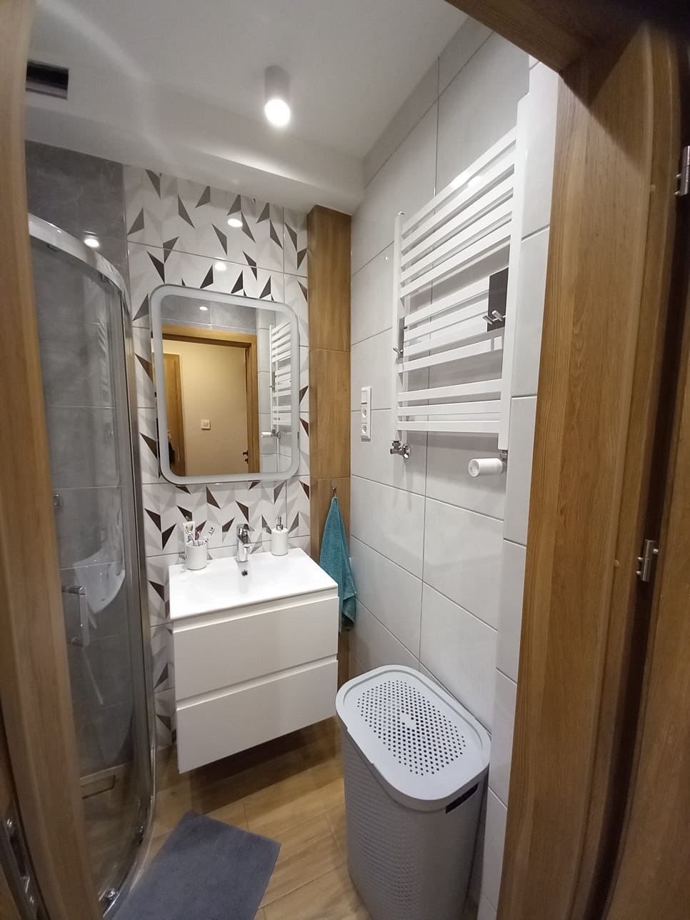 Nowoczesna łazienka z kabiną prysznicową, białą umywalką z szafką, grzejnikiem drabinkowym i koszem na pranie. Na ścianie płytki z geometrycznym wzorem w odcieniach szarości i brązu.