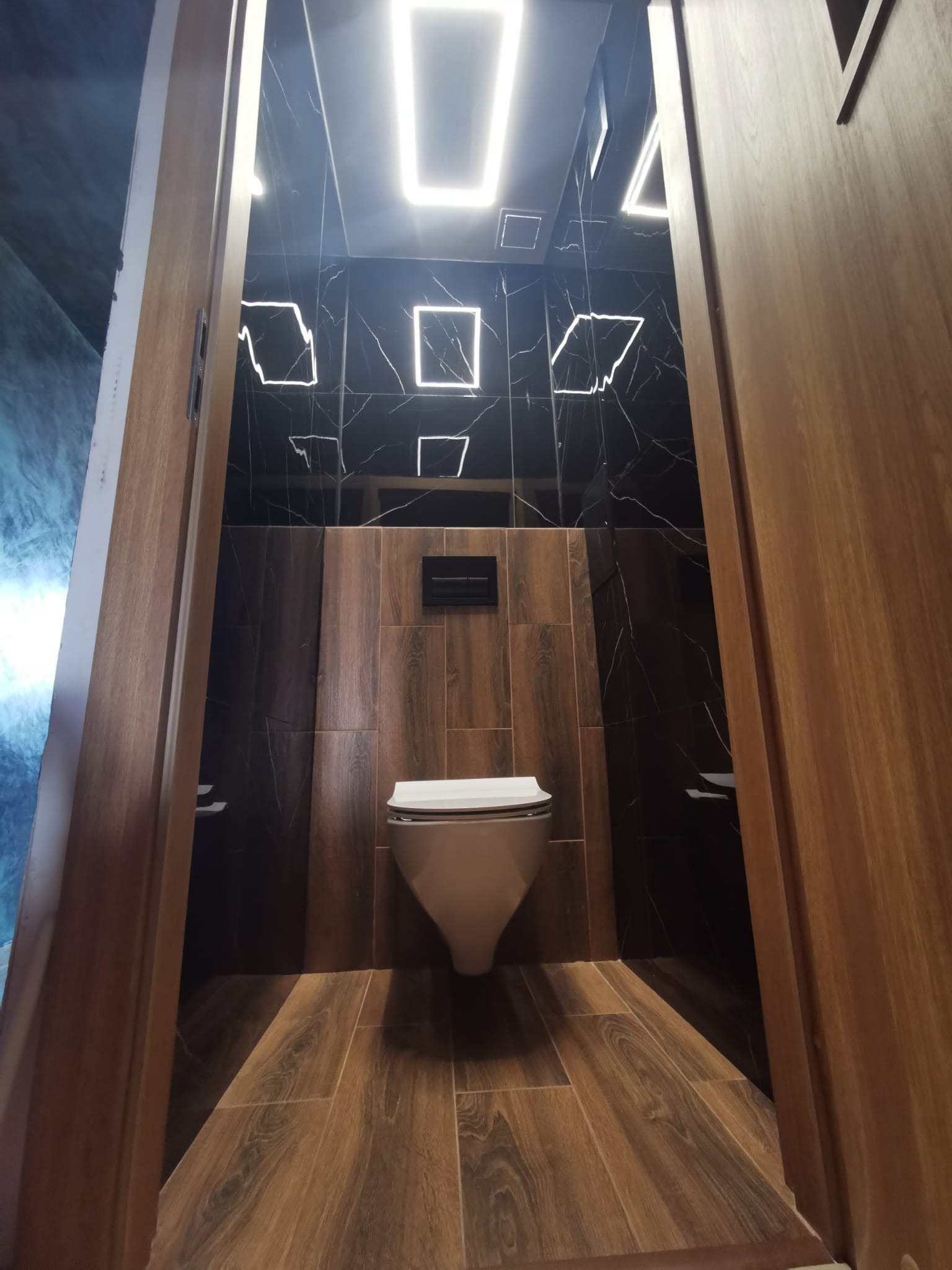 Nowoczesna toaleta z czarnymi marmurowymi płytkami i podświetlanymi kwadratami, drewnopodobna podłoga i częściowo widoczne drewniane drzwi.