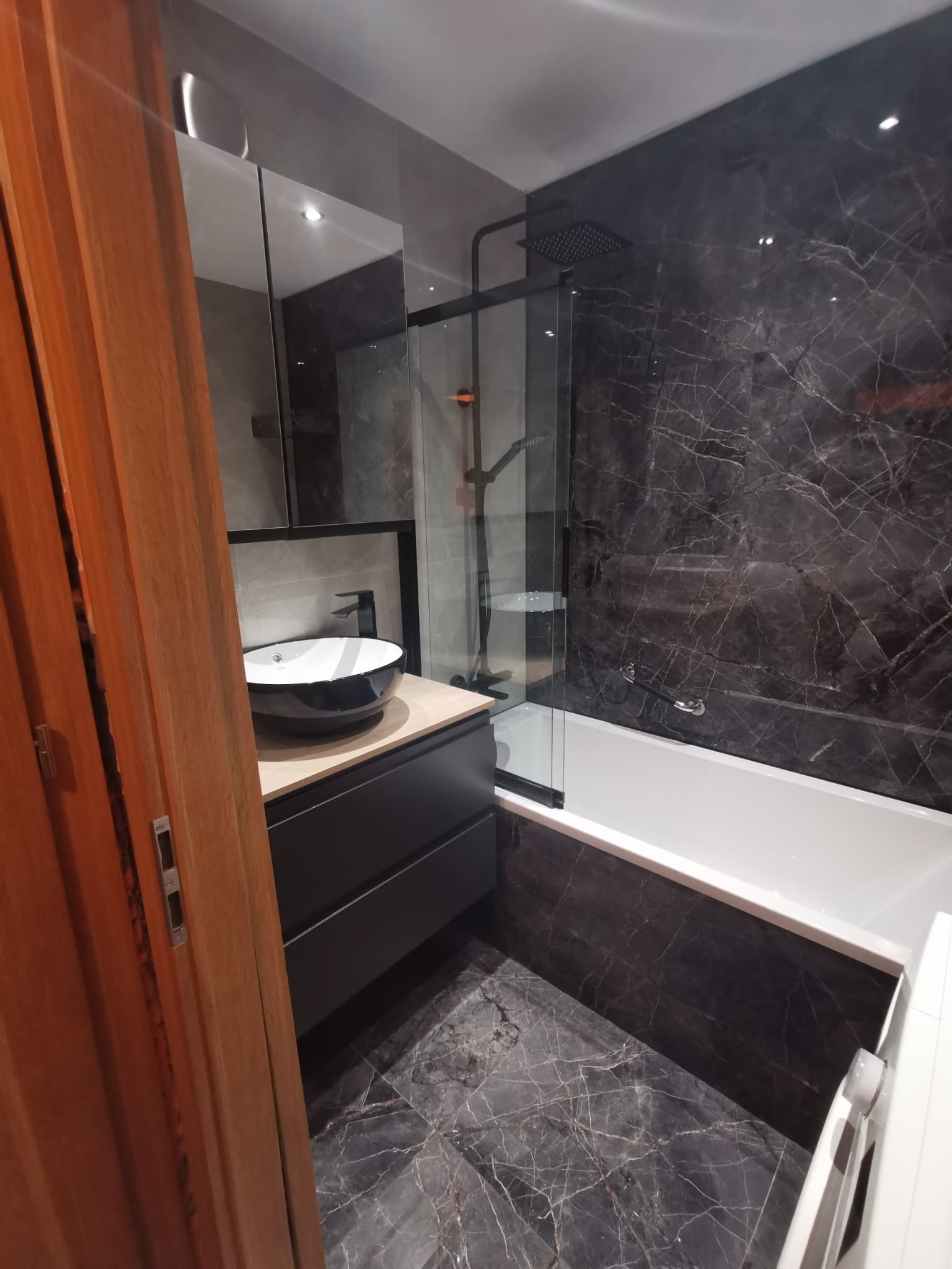 Nowoczesna łazienka z czarnymi marmurowymi płytkami, owalną umywalką na szafce z szufladami i kabiną prysznicową nad wanną, widok zza drewnianych drzwi.