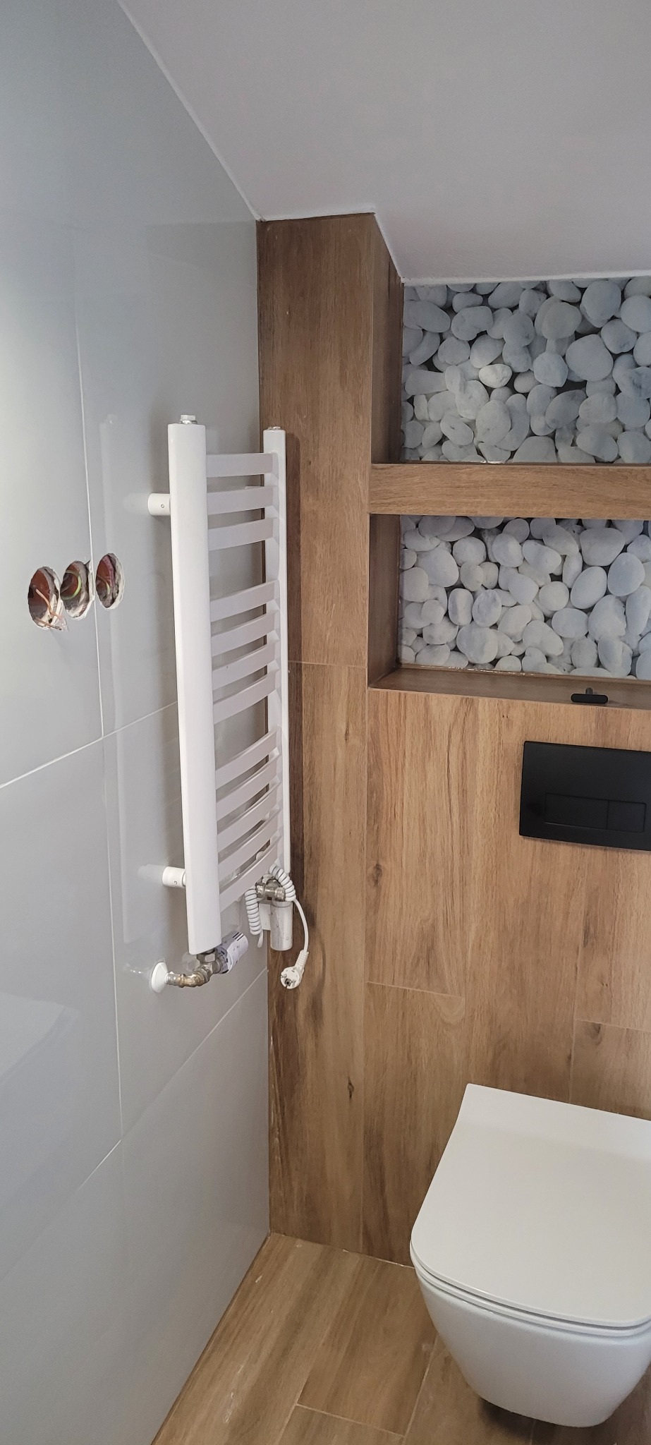 Nowoczesna łazienka w trakcie remontu: biały grzejnik drabinkowy zamontowany na szarej płytce, widoczne kable w ścianie, drewnopodobna okładzina z wnęką wyłożoną tapetą z motywem kamieni...