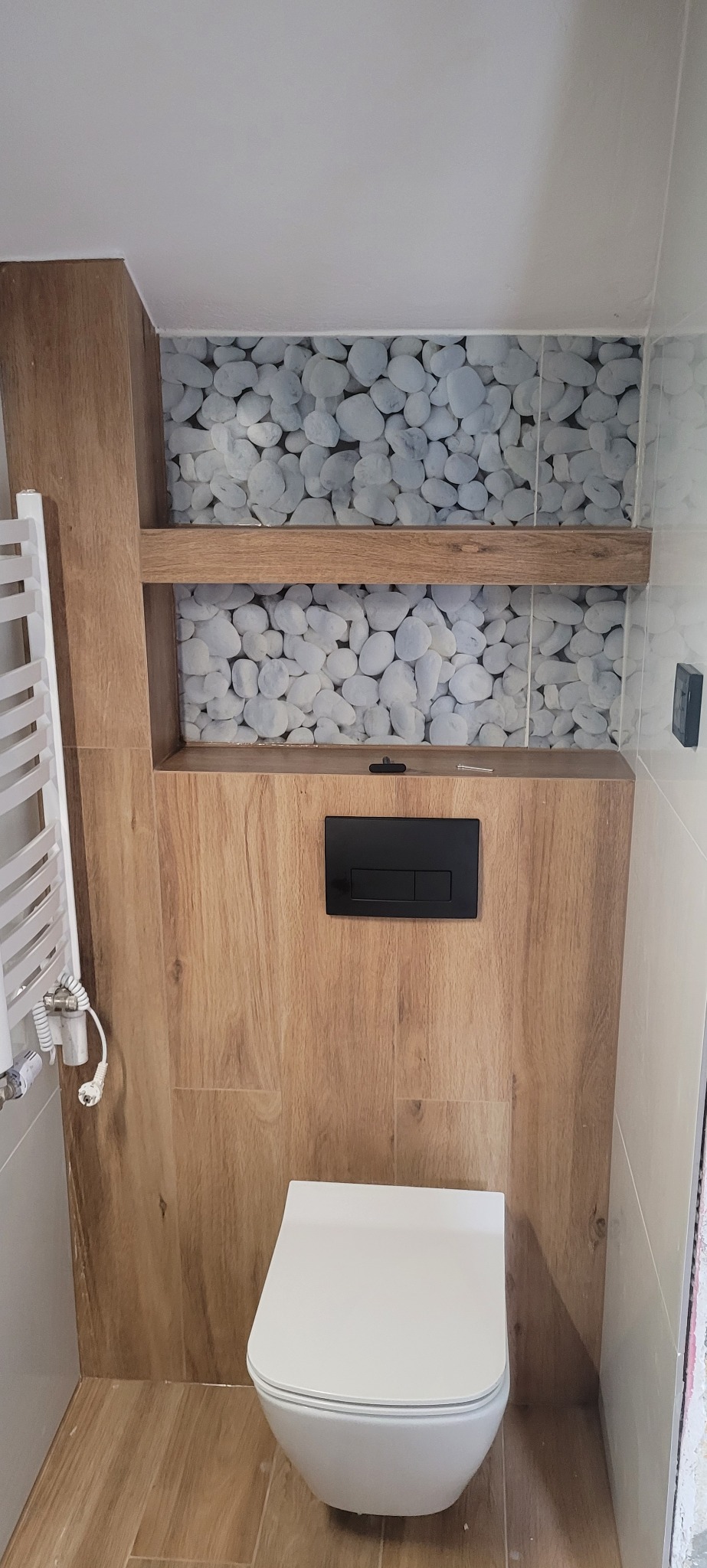 Nowoczesna toaleta podwieszana z czarnym panelem spłukującym, obudowana płytkami imitującymi drewno, z dekoracyjnym panelem z motywem białych kamieni i wnęką z półką.