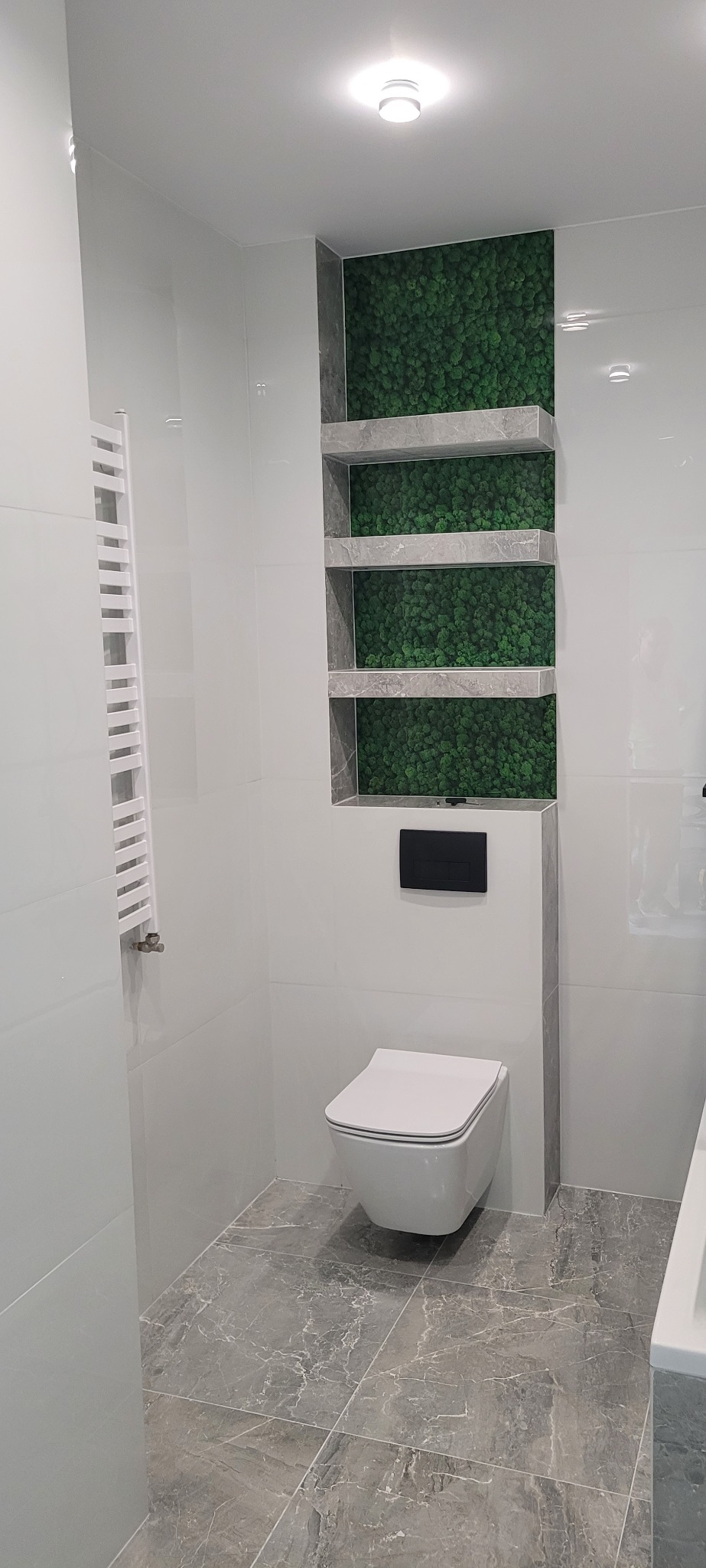 Nowoczesna łazienka z podwieszaną toaletą, szarymi płytkami podłogowymi imitującymi marmur i pionową wnęką z półkami wypełnionymi dekoracyjnym mchem.