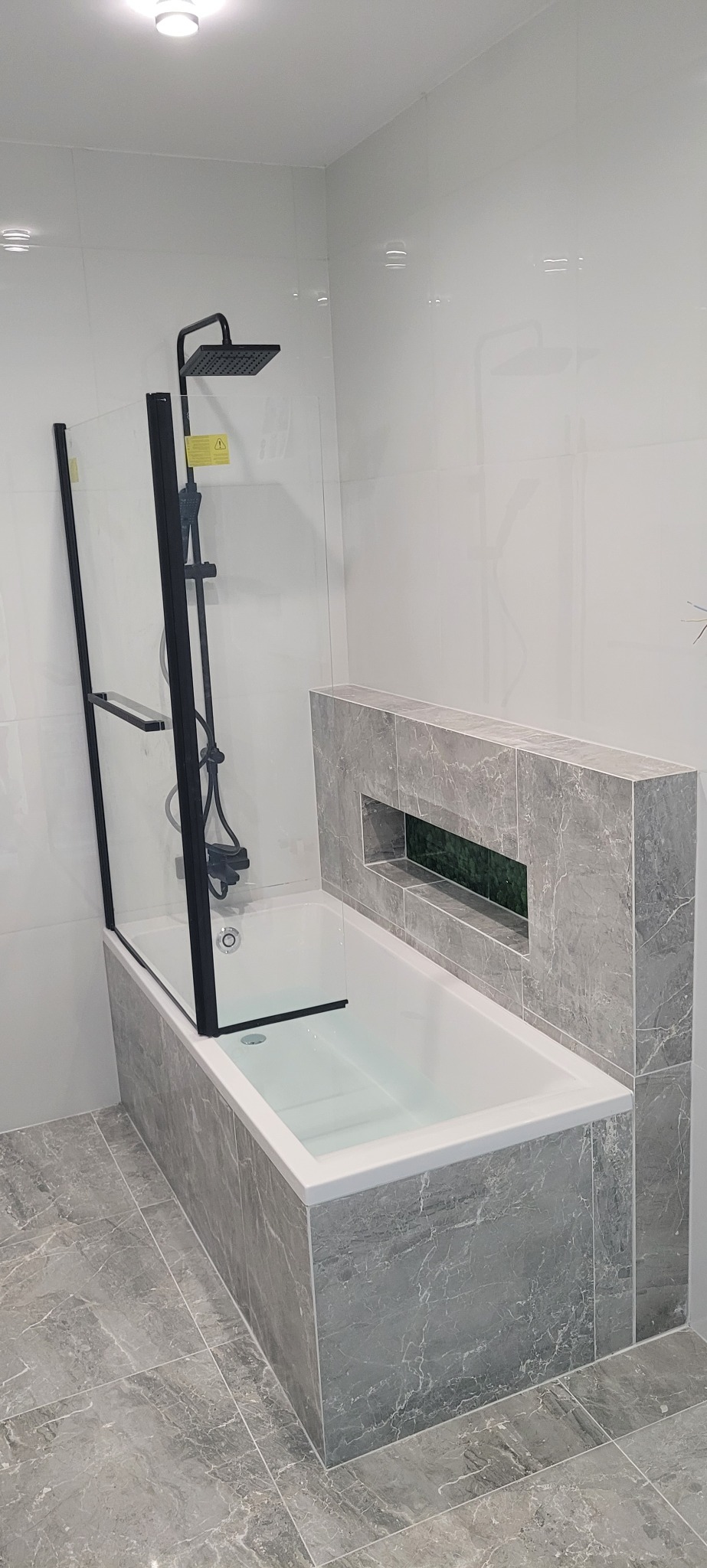 Nowoczesna łazienka z wanną obudowaną płytkami imitującymi marmur, czarną armaturą prysznicową i szklanym parawanem, wykończona w szaro-białej kolorystyce, z wnęką dekoracyjną nad wanną.