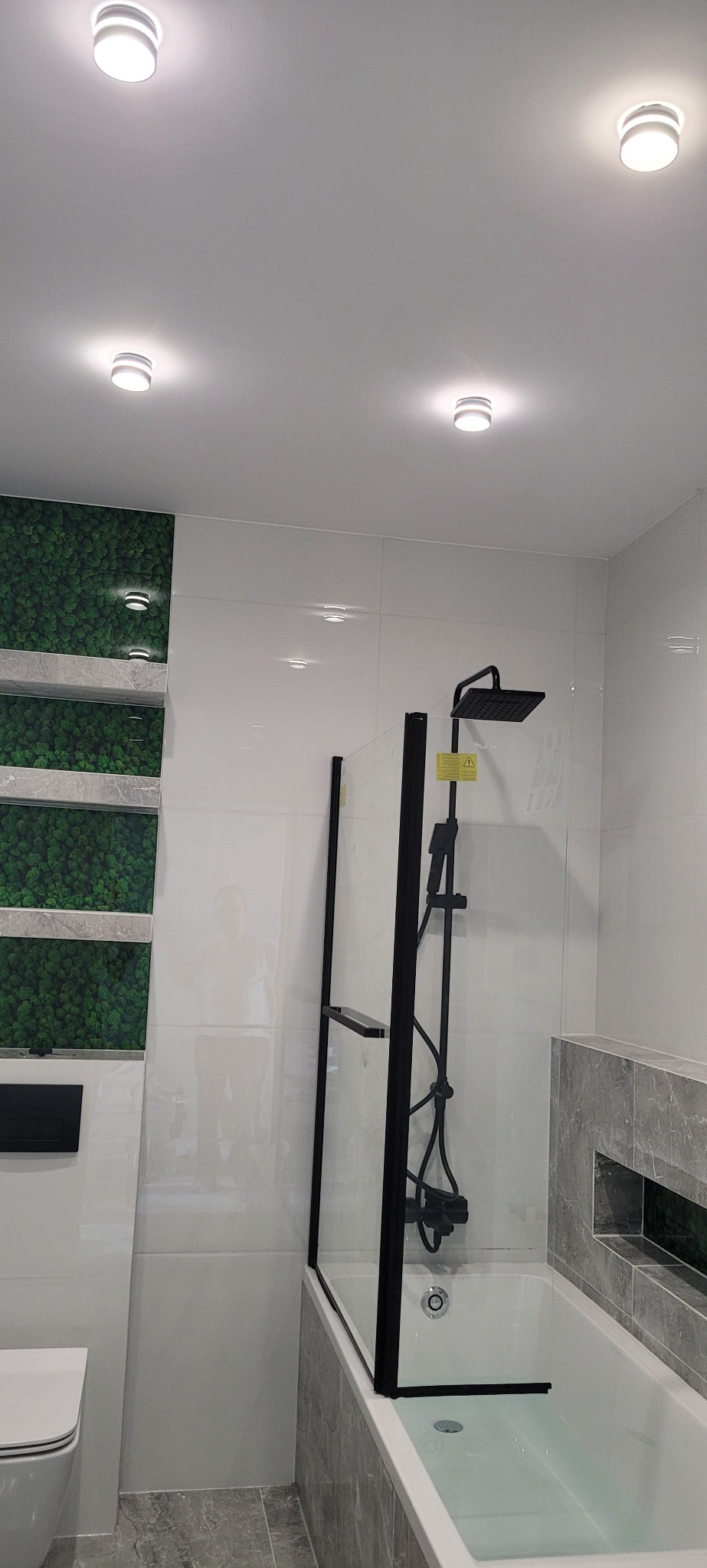 Nowoczesna łazienka z wanną obudowaną szarymi płytkami imitującymi marmur, czarną armaturą, szklanym parawanem nawannowym z czarną ramą oraz panelem z zielonego mchu.