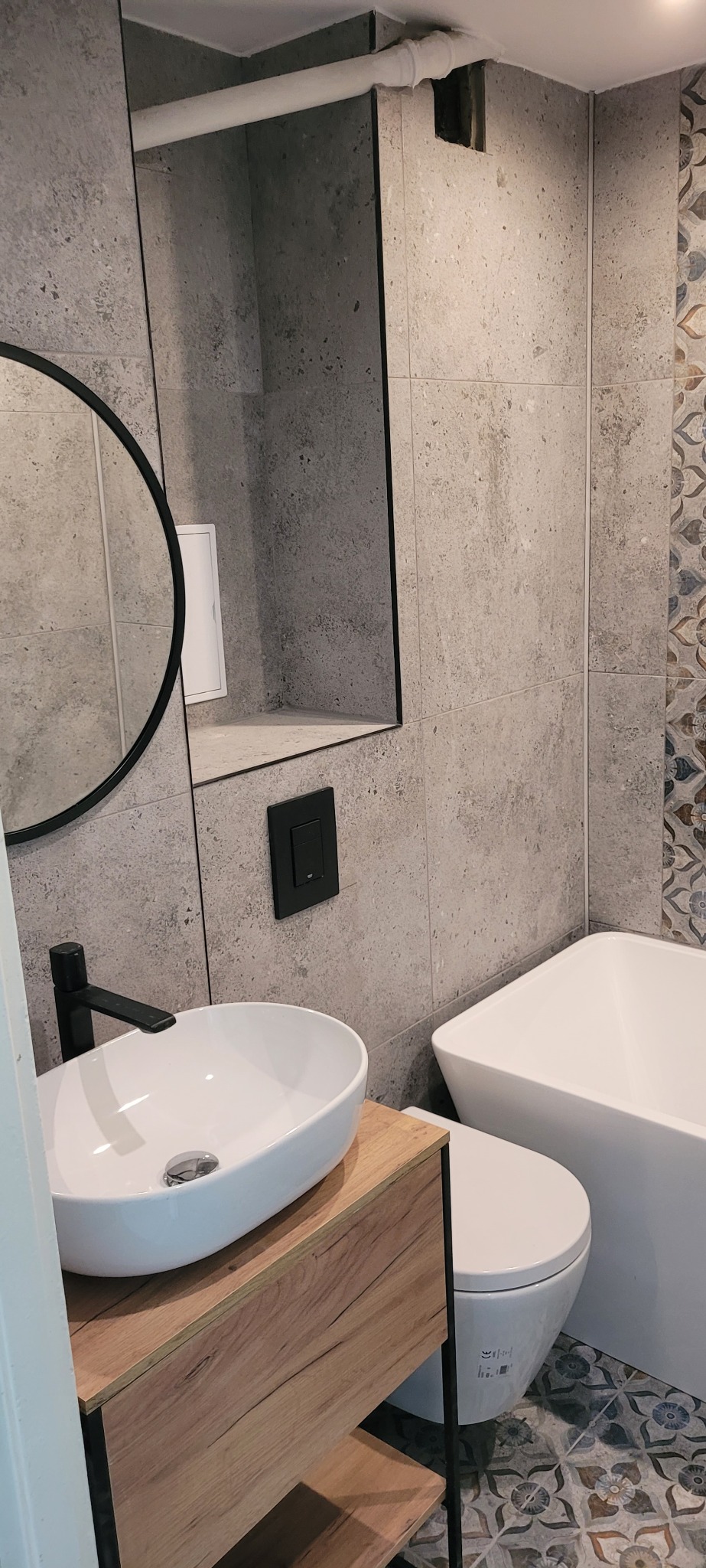 Nowoczesna łazienka z okrągłym lustrem, umywalką nablatową na drewnianej szafce, białą wanną i toaletą, wykończona szarymi płytkami imitującymi beton i dekoracyjnymi płytkami na ścianie.