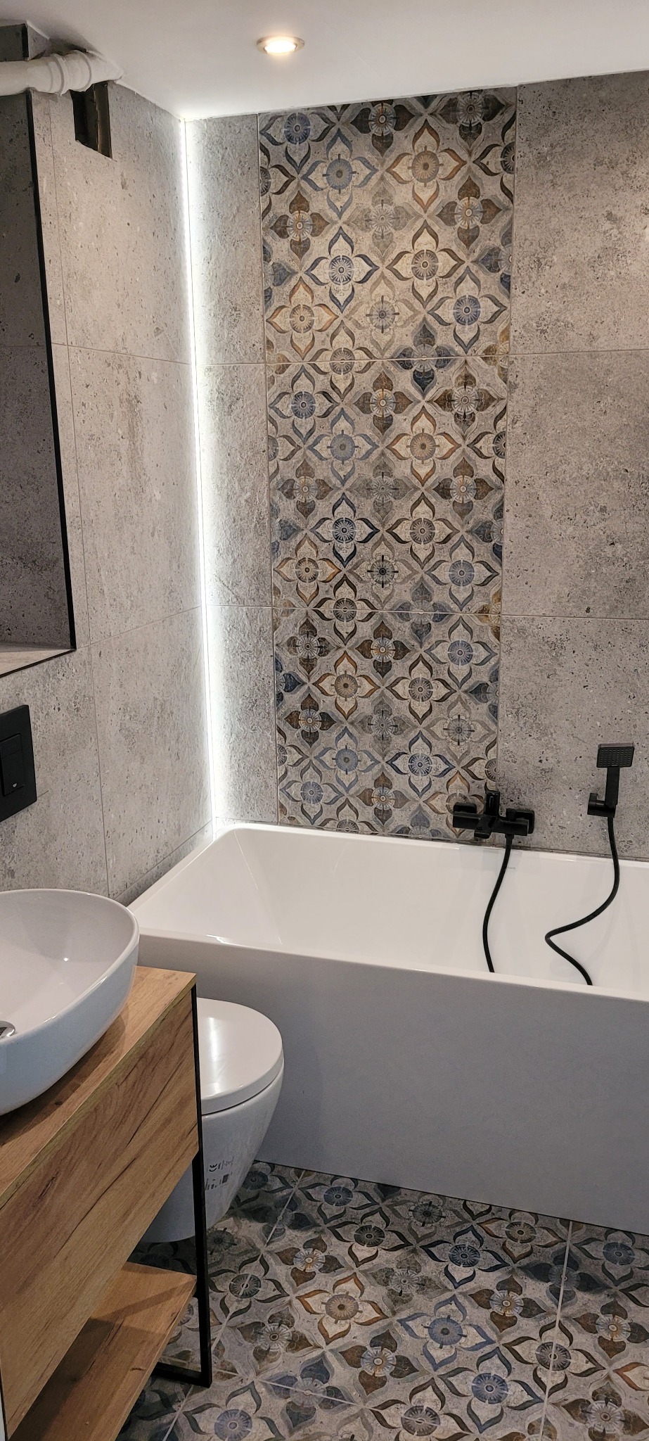 Nowoczesna łazienka z wanną, umywalką na drewnianej szafce i toaletą; ściany wykończone szarymi płytkami i dekoracyjnym pasem płytek z motywem kwiatowym, podłoga z podobnym wzorem; czarna armatura.