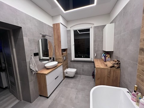 Wykończona łazienka w szarościach z drewnianymi akcentami, owalna umywalka na białej szafce z szufladami, okrągłe lustro, płytki imitujące beton na ścianach i podłodze, sufit z podświetleniem LED.