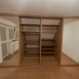 Jasna, drewniana szafa wnękowa z lustrami po bokach, ukazująca puste półki i drążki na ubrania. Widoczna podłoga z paneli i odbicie pokoju w lustrach.