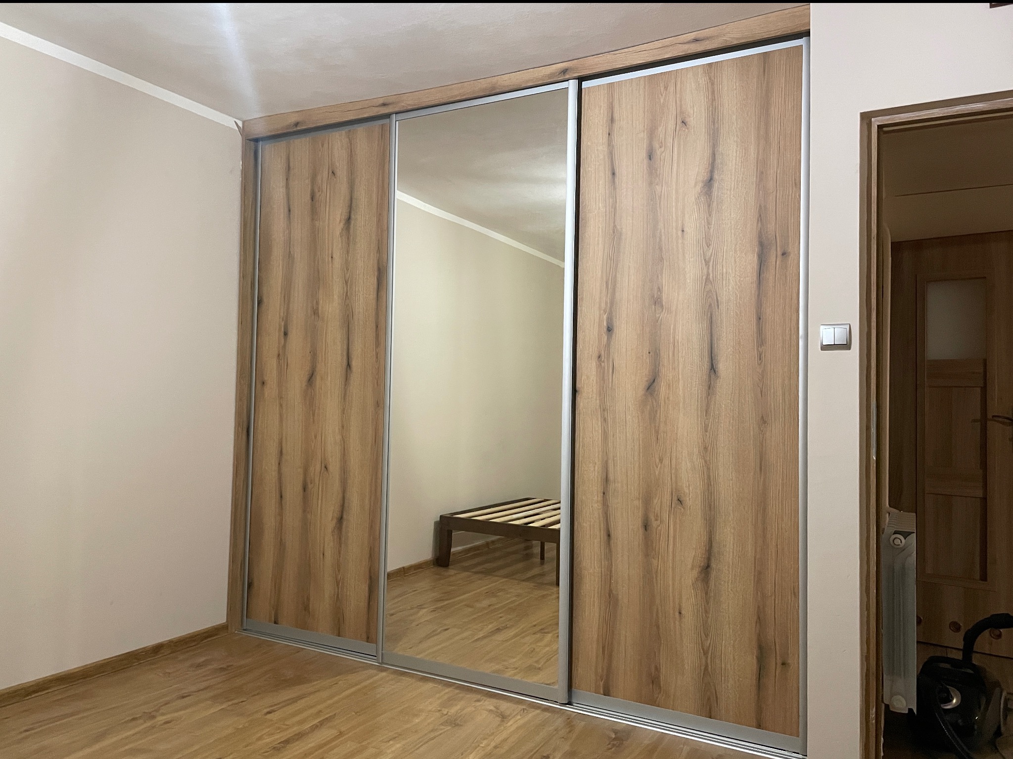 Trzydrzwiowa szafa przesuwna z lustrem, wykończona płytą meblową w odcieniu dębu, z widocznymi okuciami w kolorze naturalnego aluminium, odbijająca w lustrze ramę łóżka na drewnianej podłodze.