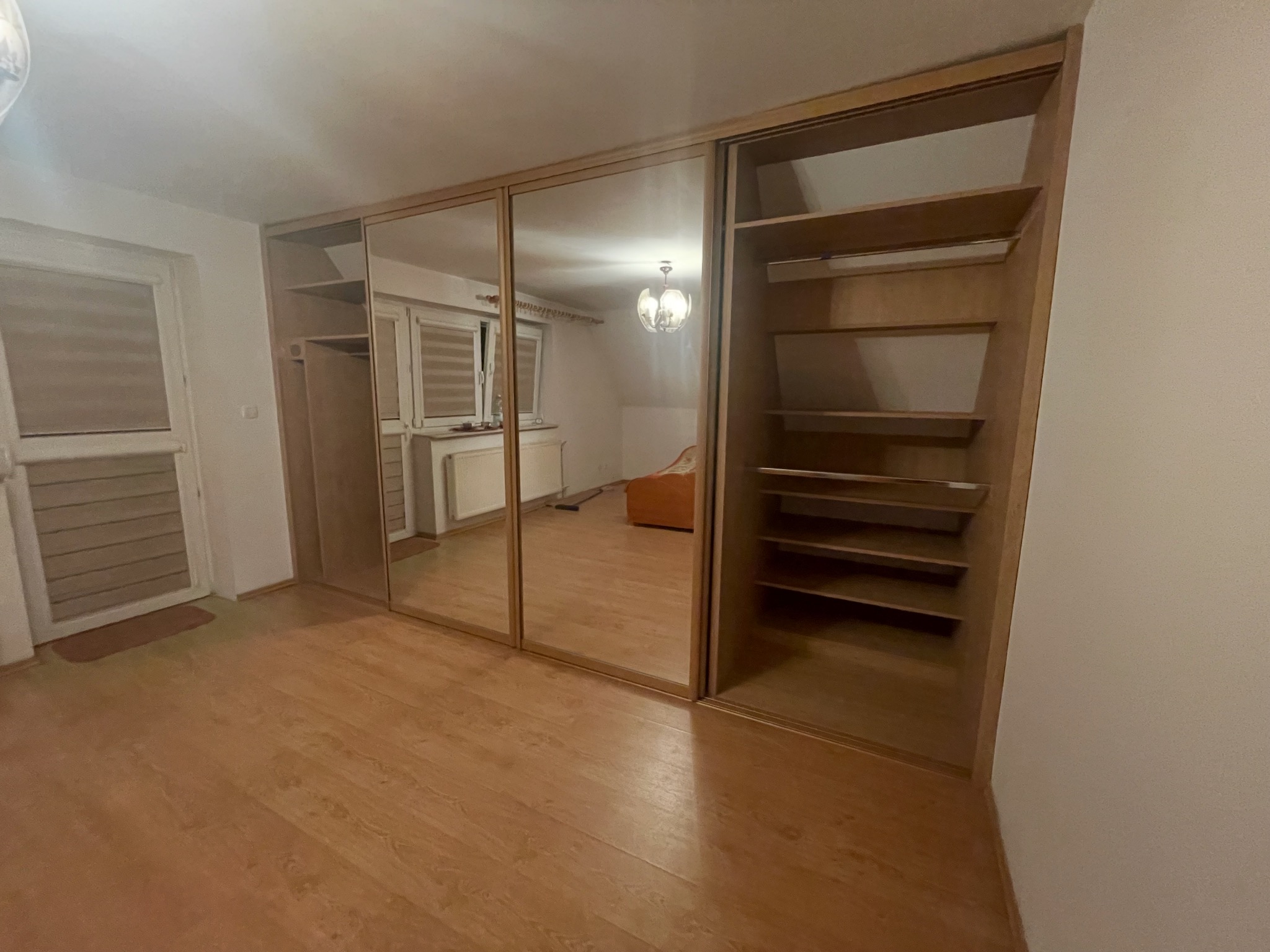 Jasna szafa wnękowa z lustrzanymi drzwiami przesuwnymi i otwartymi półkami, widoczna na tle pokoju z panelami podłogowymi. Szafa z drewnianymi elementami.