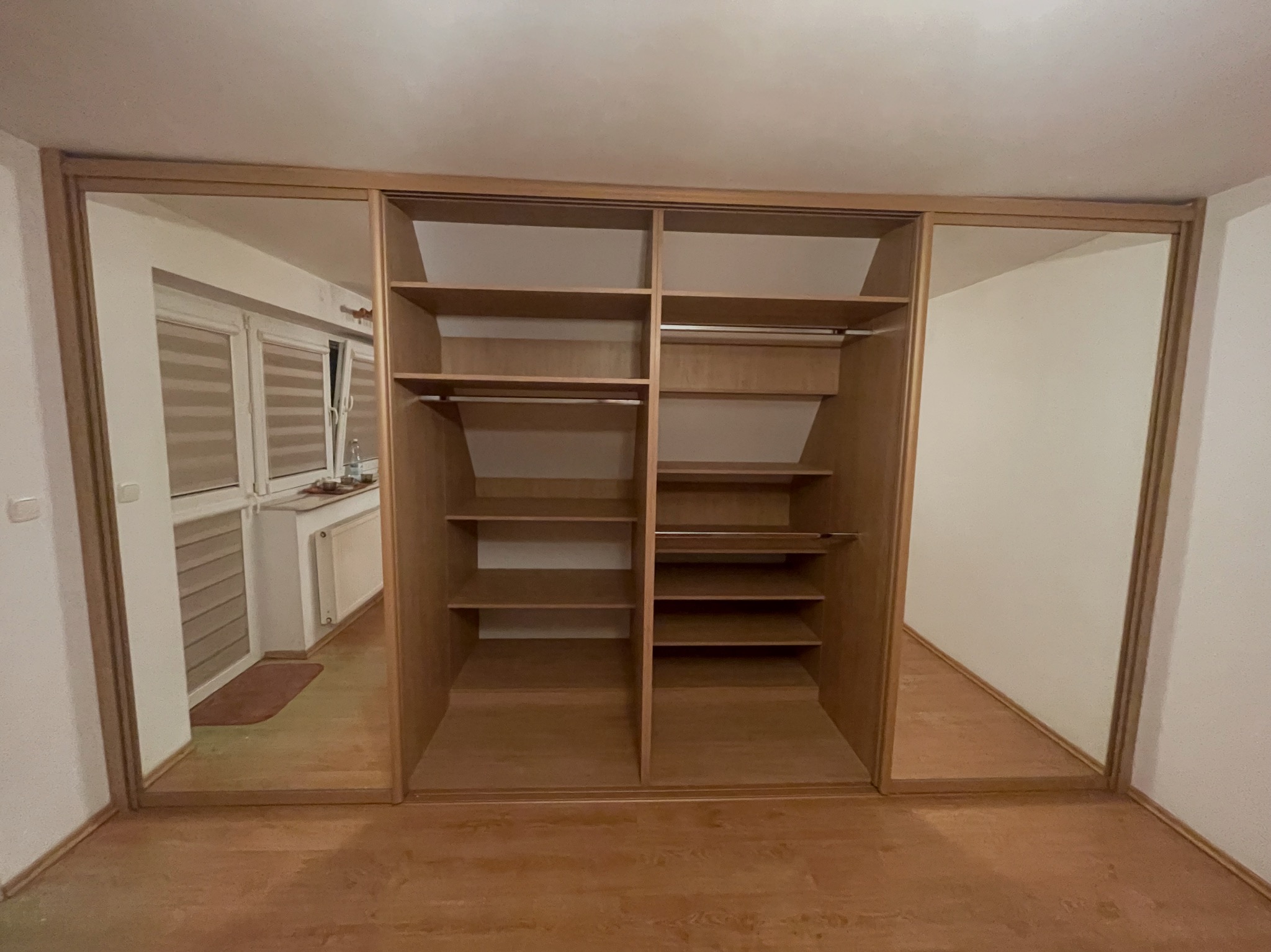 Jasna, drewniana szafa wnękowa z lustrami po bokach, ukazująca puste półki i drążki na ubrania. Widoczna podłoga z paneli i odbicie pokoju w lustrach.