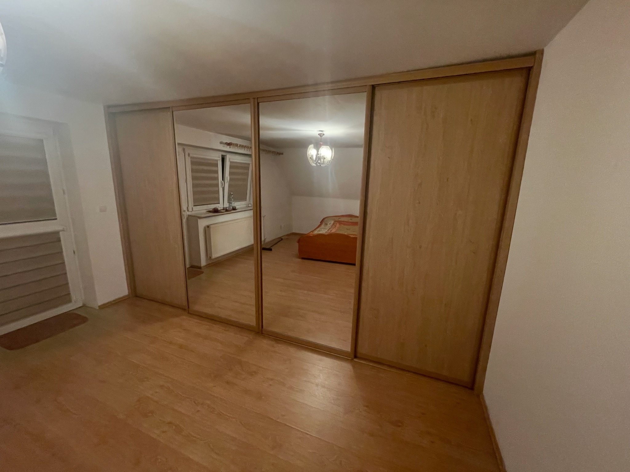 Drewniana szafa wnękowa z lustrzanymi drzwiami przesuwnymi w Bałtowie, odbijająca wnętrze pokoju. Widoczne panele podłogowe i fragment okna.