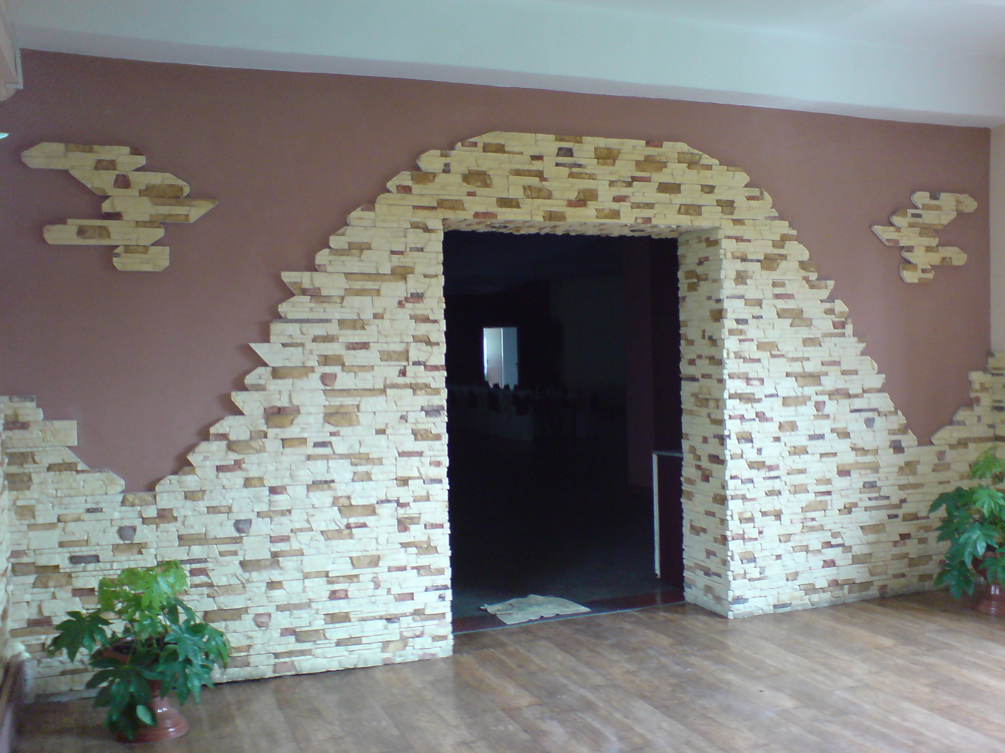 Wykończenie ściany wokół drzwi imitacją kamienia w Katowicach, z dekoracyjnymi elementami i roślinami doniczkowymi.