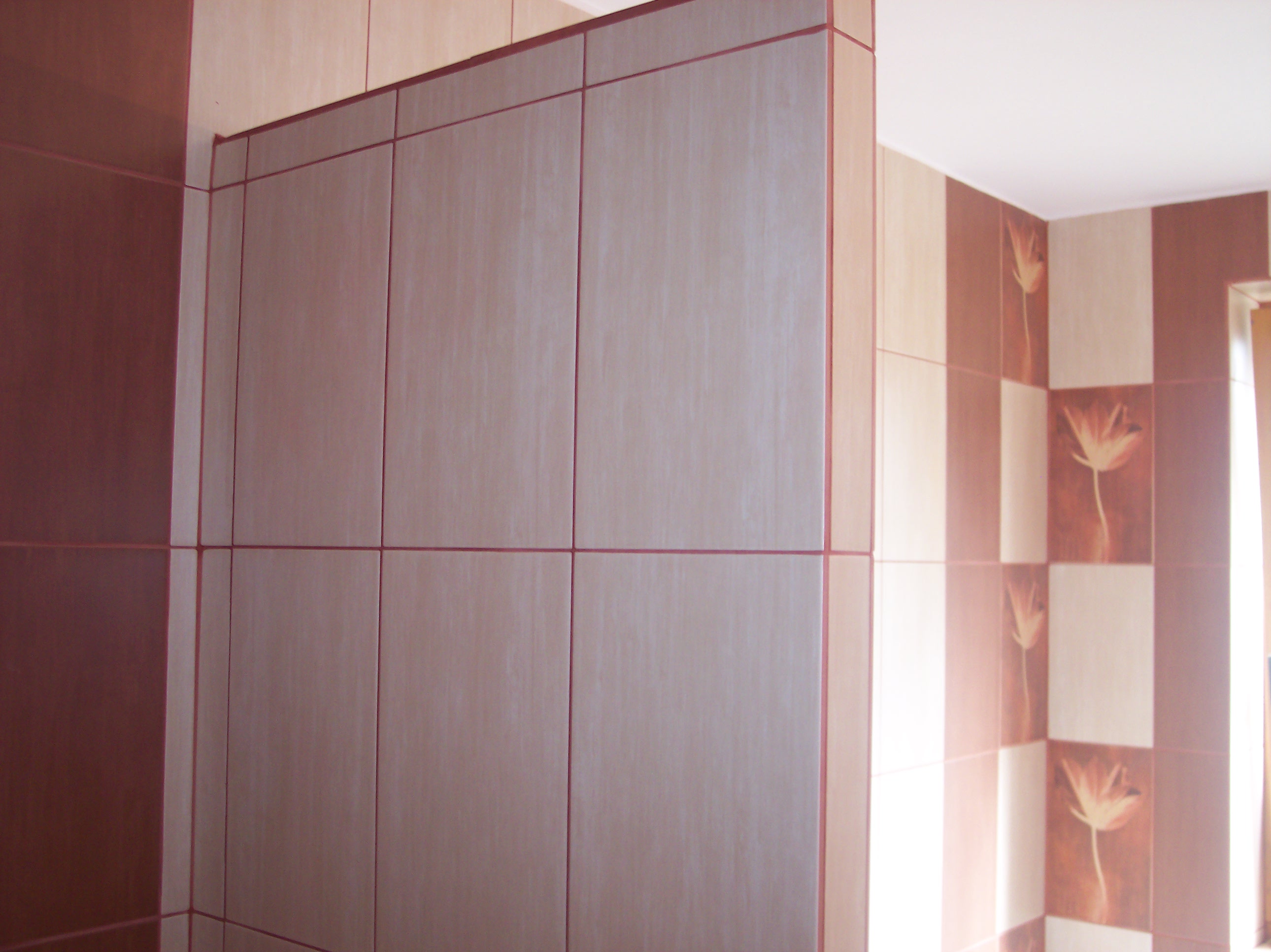 Wykończona ściana w łazience z szarych płytek ceramicznych z bordową fugą i dekoracyjnymi płytkami z motywem kwiatowym na sąsiedniej ścianie.