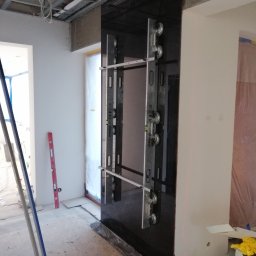 8 Piętro Sp. z o.o. - Montaż czarnej, błyszczącej płyty wielkoformatowej na ścianie, przy użyciu przyssawek i poziomicy. Widoczne narzędzia glazurnicze i surowy stan wykończenia wnętrza.