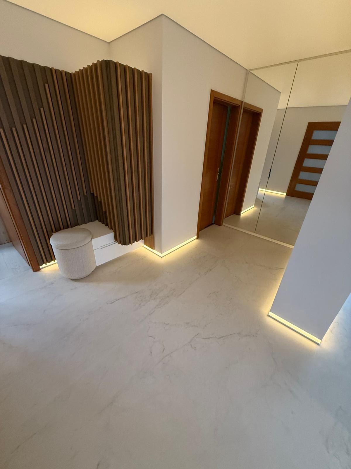 Nowoczesny przedpokój z drewnianą ścianką dekoracyjną, podświetleniem LED i marmurową posadzką. Lustro na ścianie powiększa optycznie przestrzeń.