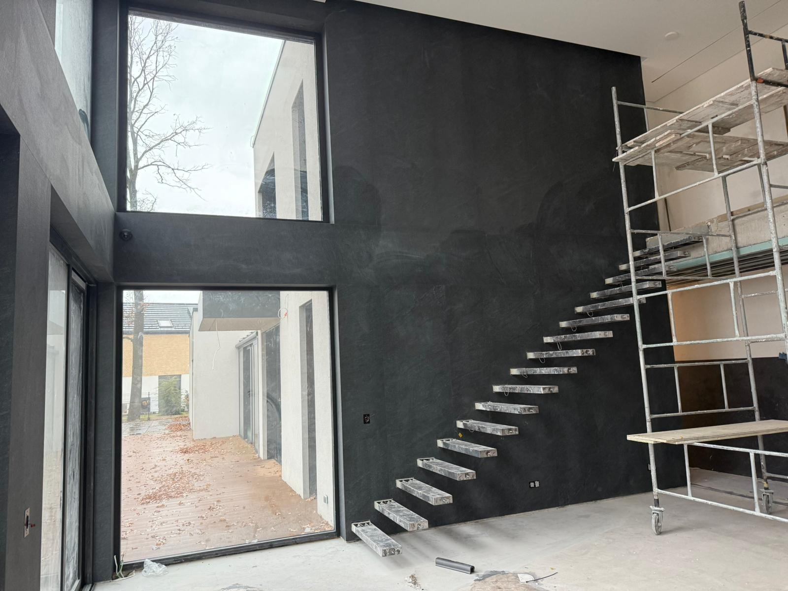 Nowoczesne wnętrze w trakcie wykończenia: czarna ściana z fakturą, minimalistyczne schody i rusztowanie. Duże okna z widokiem na zewnątrz.